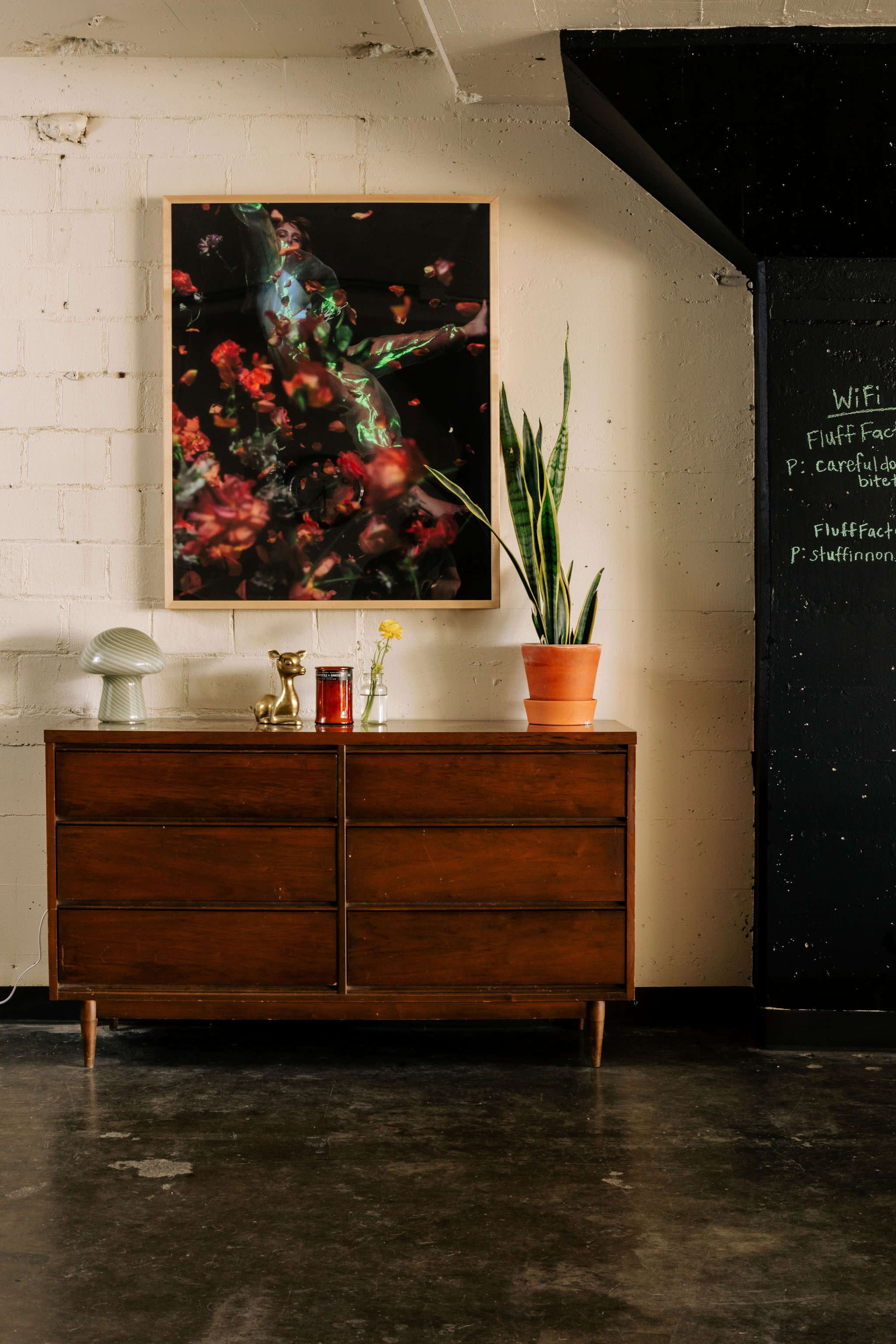 A wooden dresser with drawers displays a potted plant and a vase of flowers in front of a wall featuring a large abstract painting and a chalkboard.