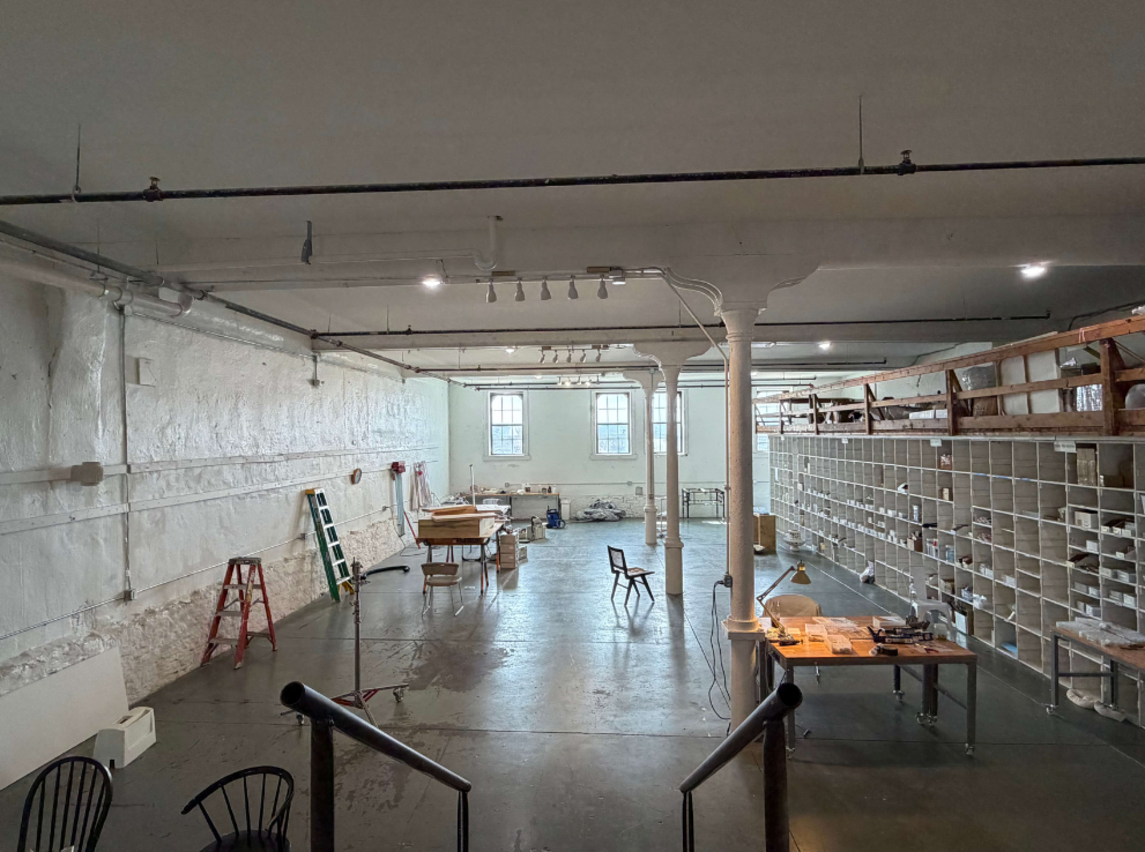The image shows a spacious industrial-style room with white walls, wooden shelving, and various worktables scattered throughout.