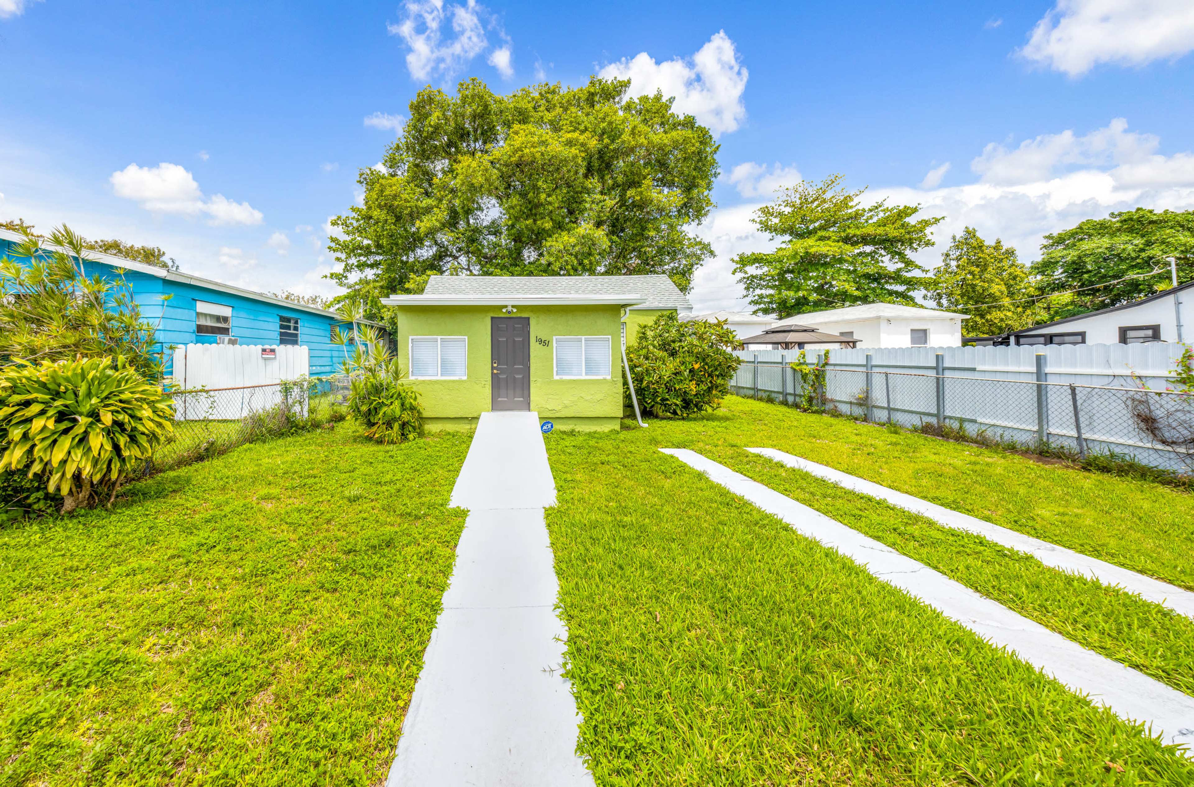 The image shows a small green house with a gray roof, set on a grassy yard with a concrete walkway leading to the entrance, surrounded by trees and nearby buildings.
