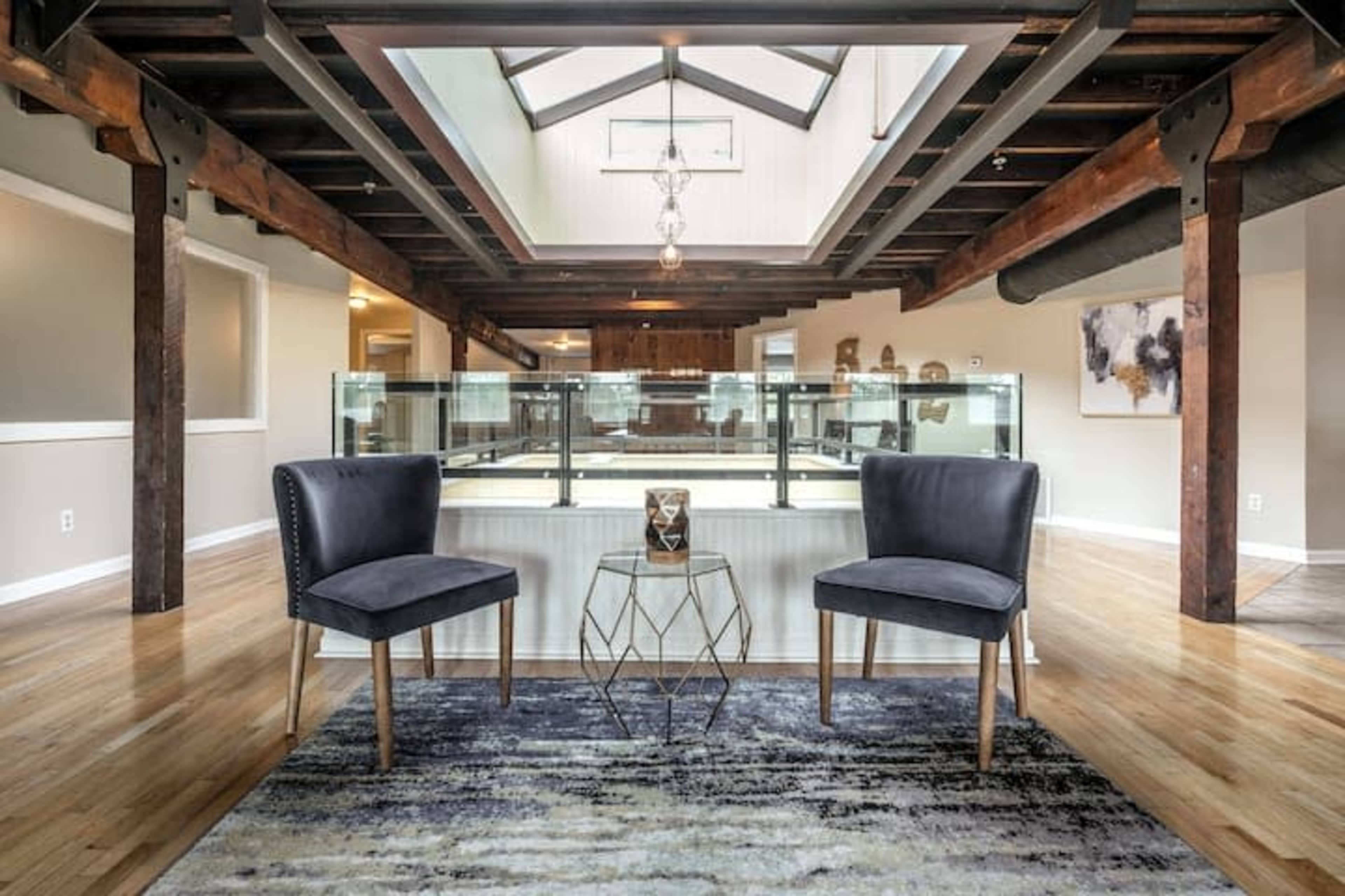 A modern interior space featuring two gray chairs on a rug, a small table between them, and a large illuminated area with wooden beams and a staircase in the background.