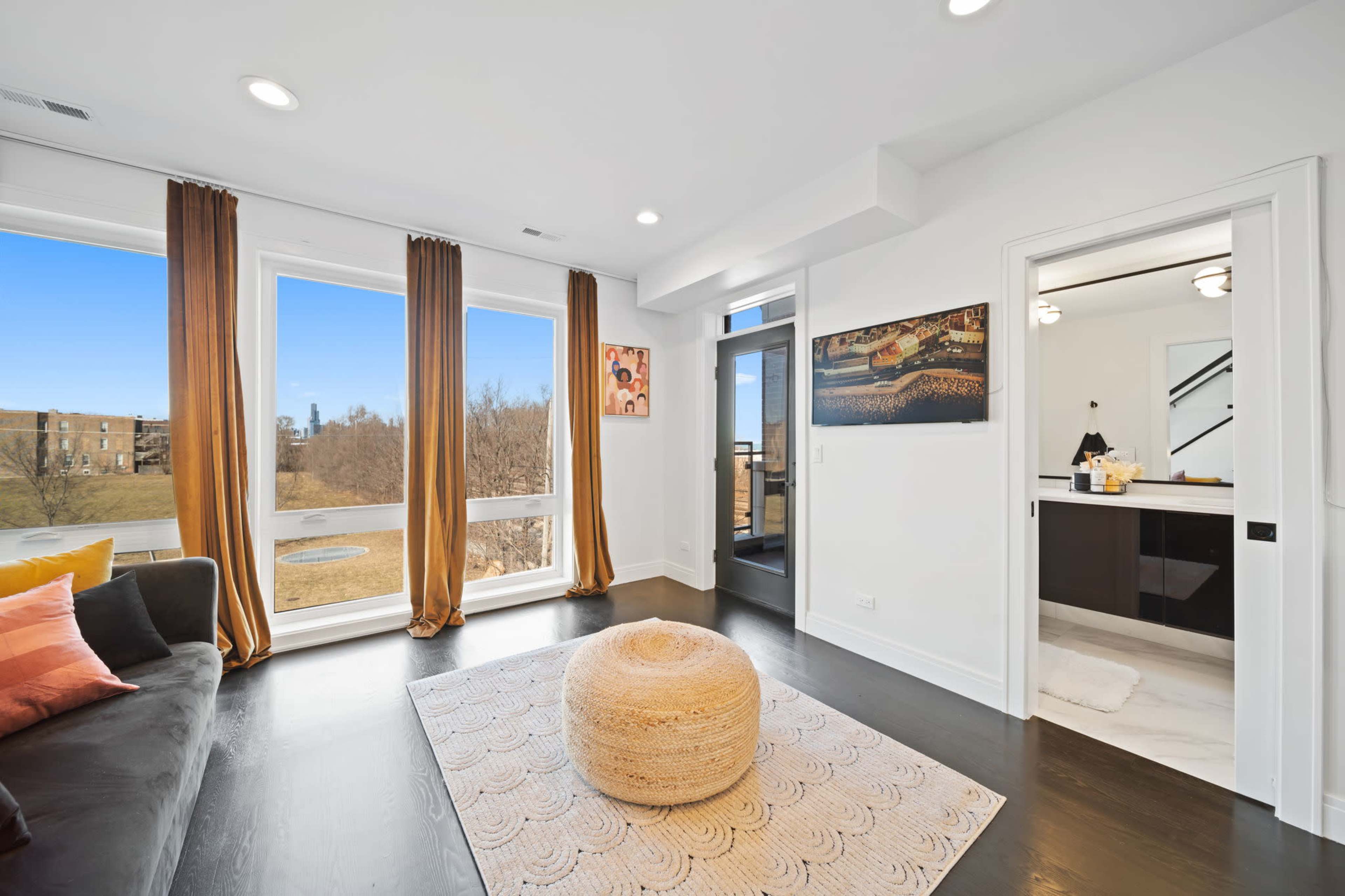 A modern living room features large windows with light curtains, a sofa, a circular ottoman, a decorative rug, and a view of a landscape outside.