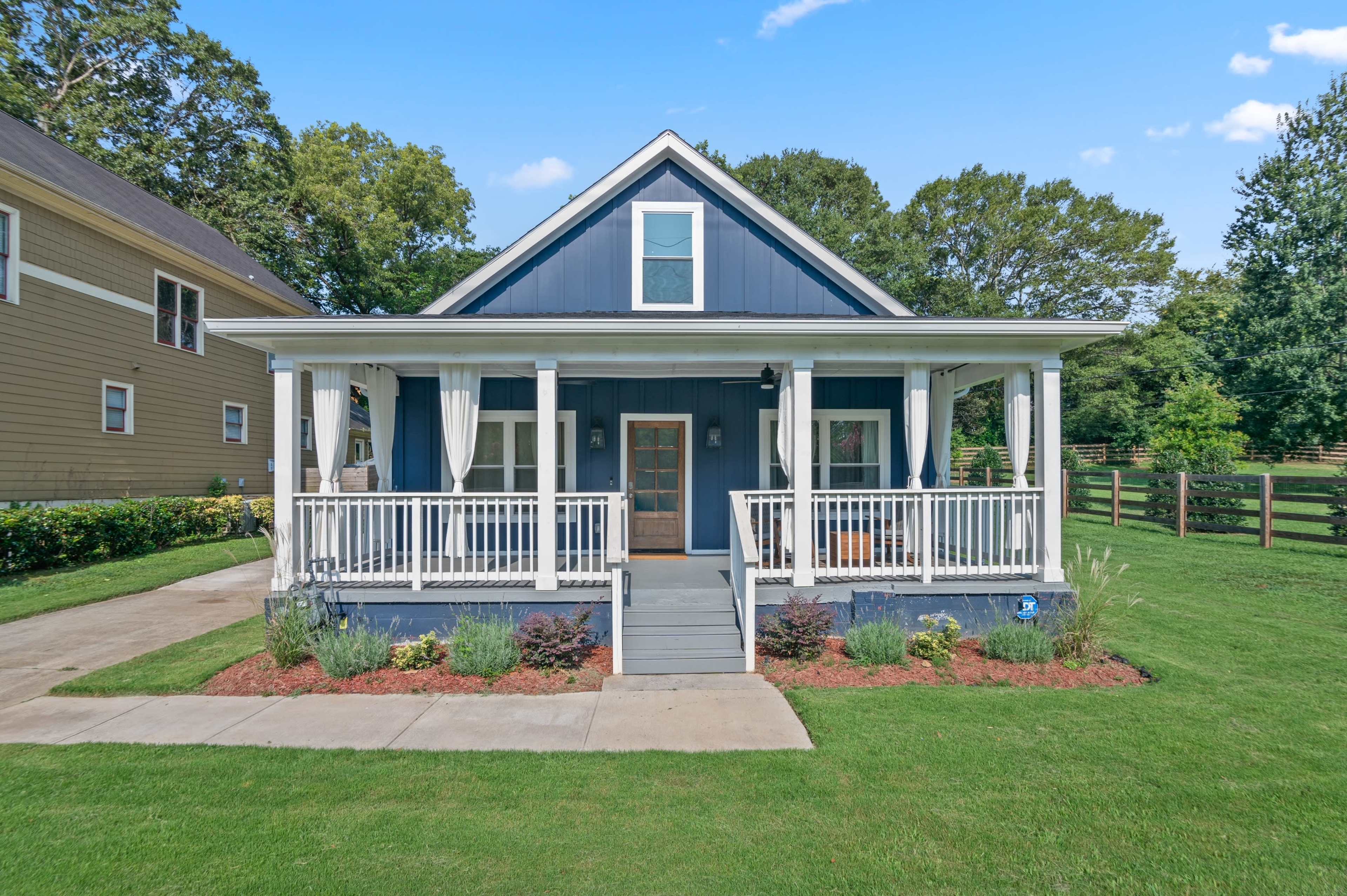 The image shows a blue cottage-style house with a front porch, white railings, and landscaped greenery in the yard.
