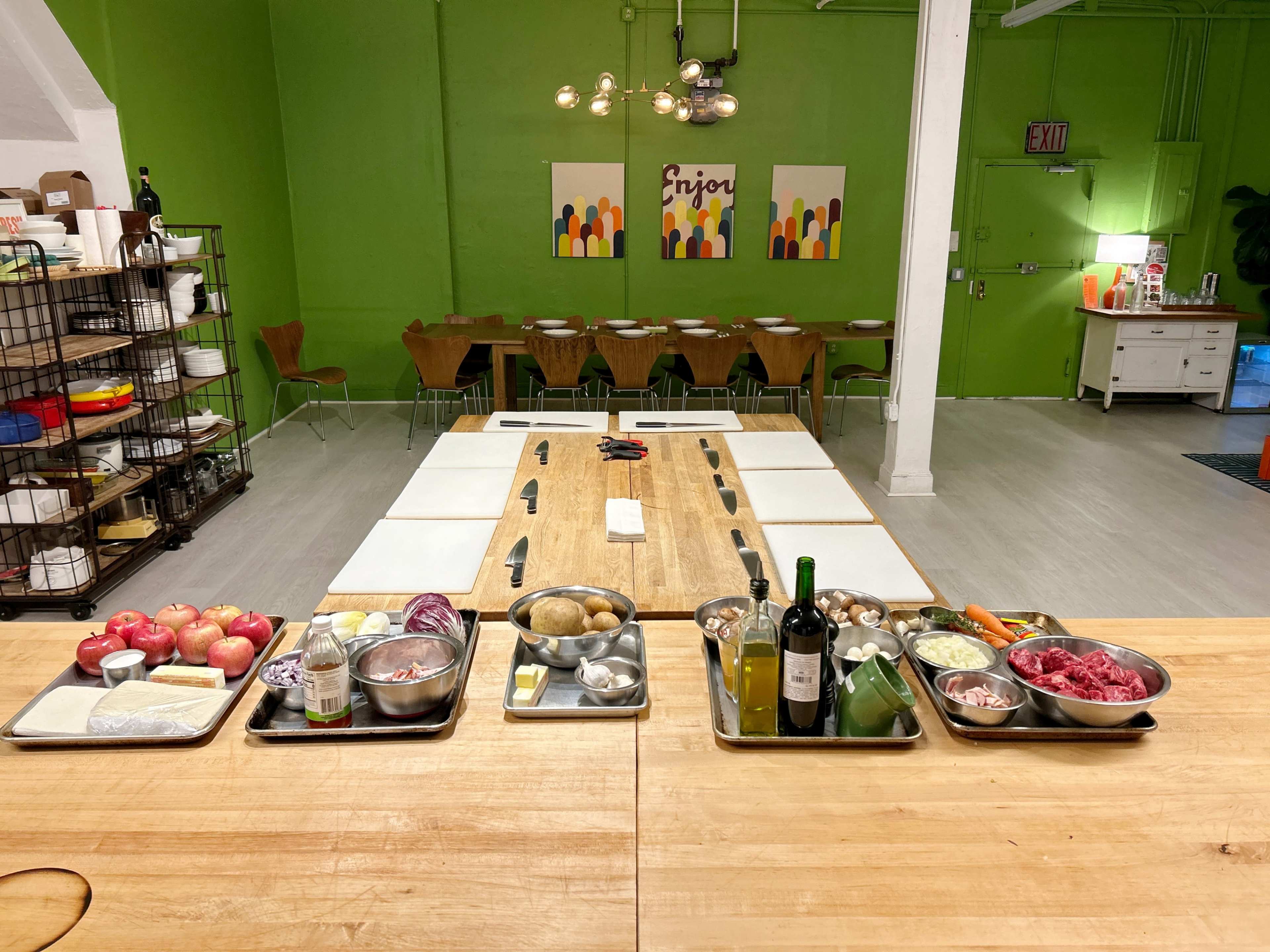 A spacious kitchen area features a large wooden table surrounded by chairs, with various food ingredients organized on trays along one side.