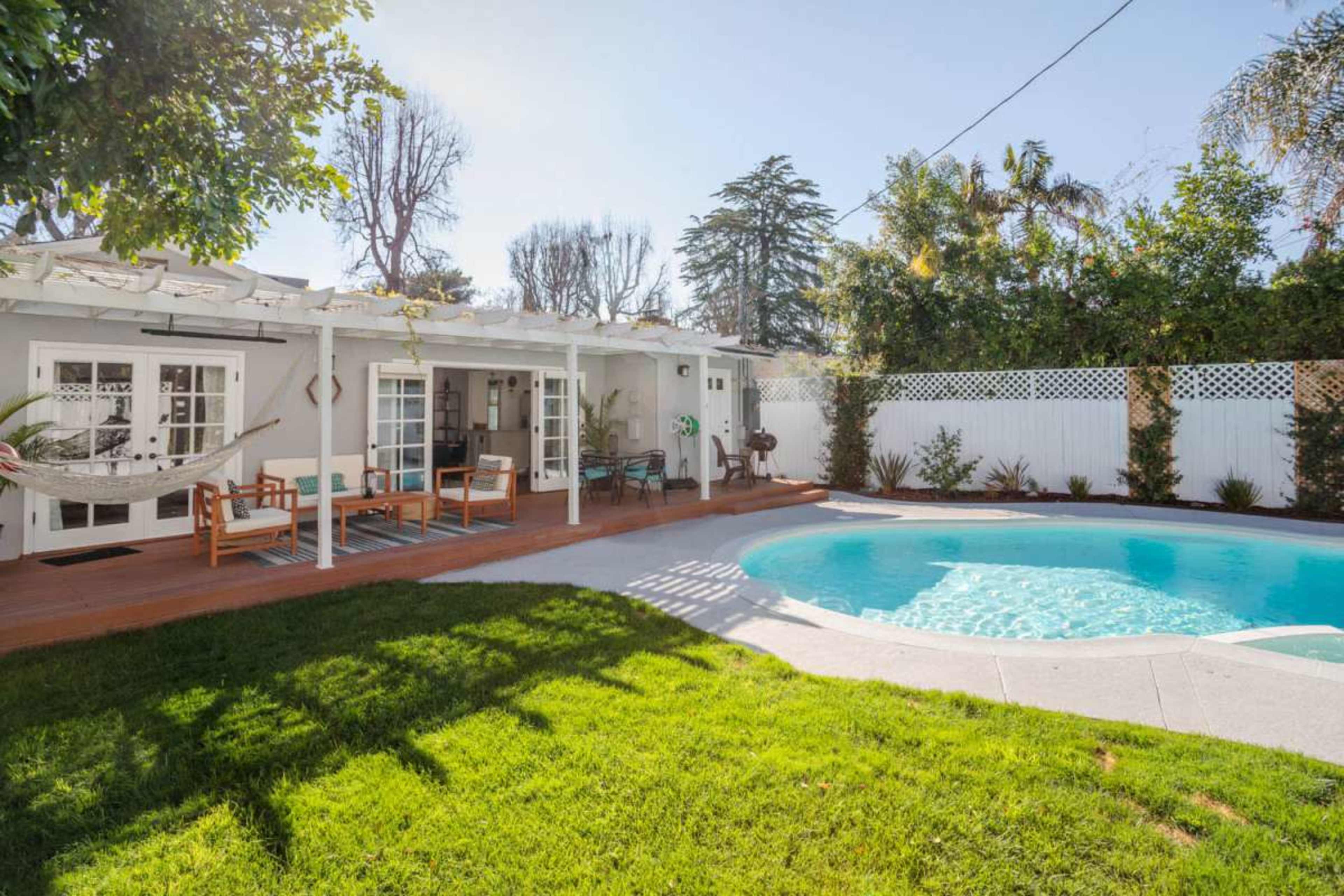 The image shows a backyard with a swimming pool, patio furniture, and a white fence surrounded by greenery.