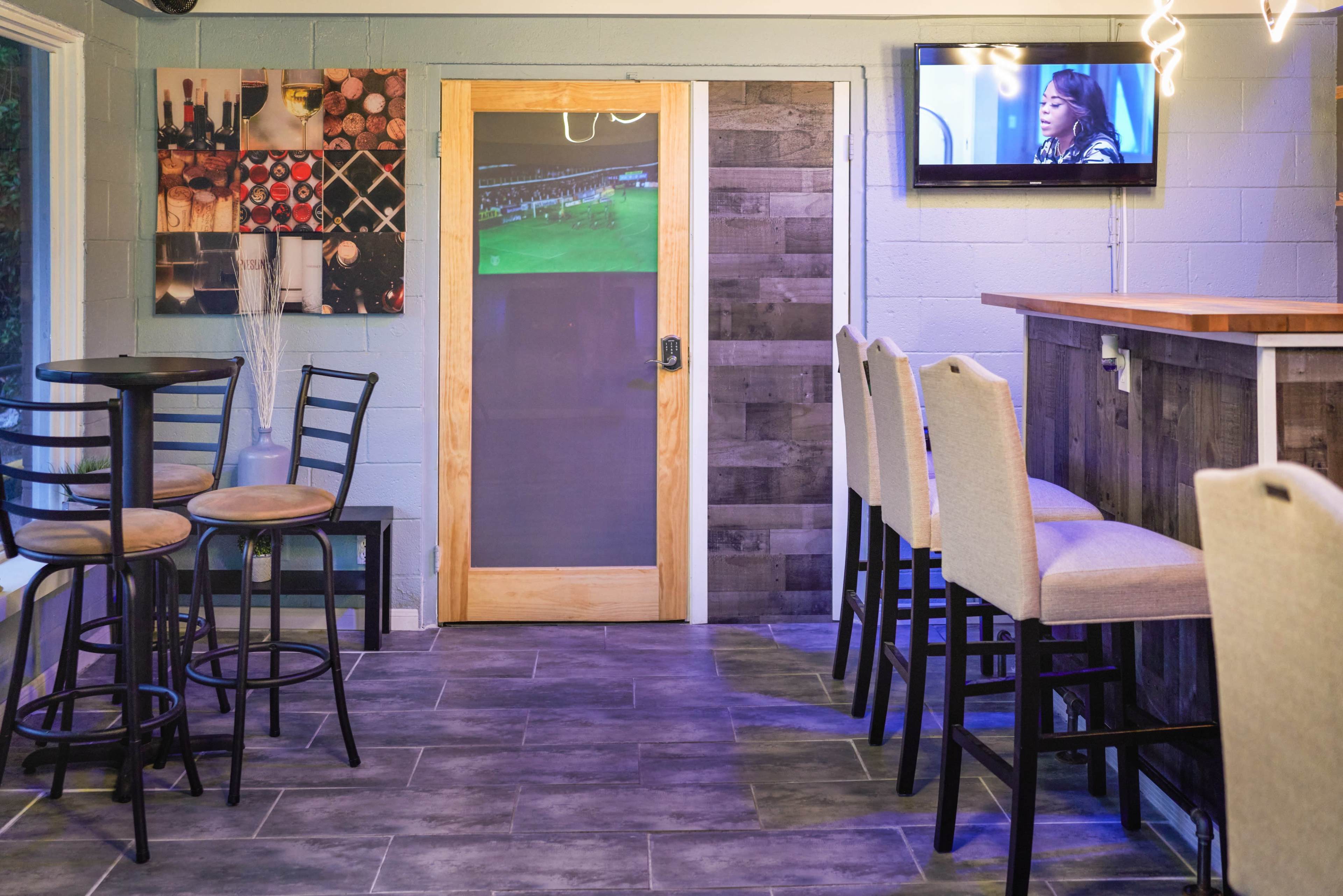 A small restaurant or bar interior with a combination of high-top and regular seating, a wooden door, and a television mounted on the wall displaying a program.