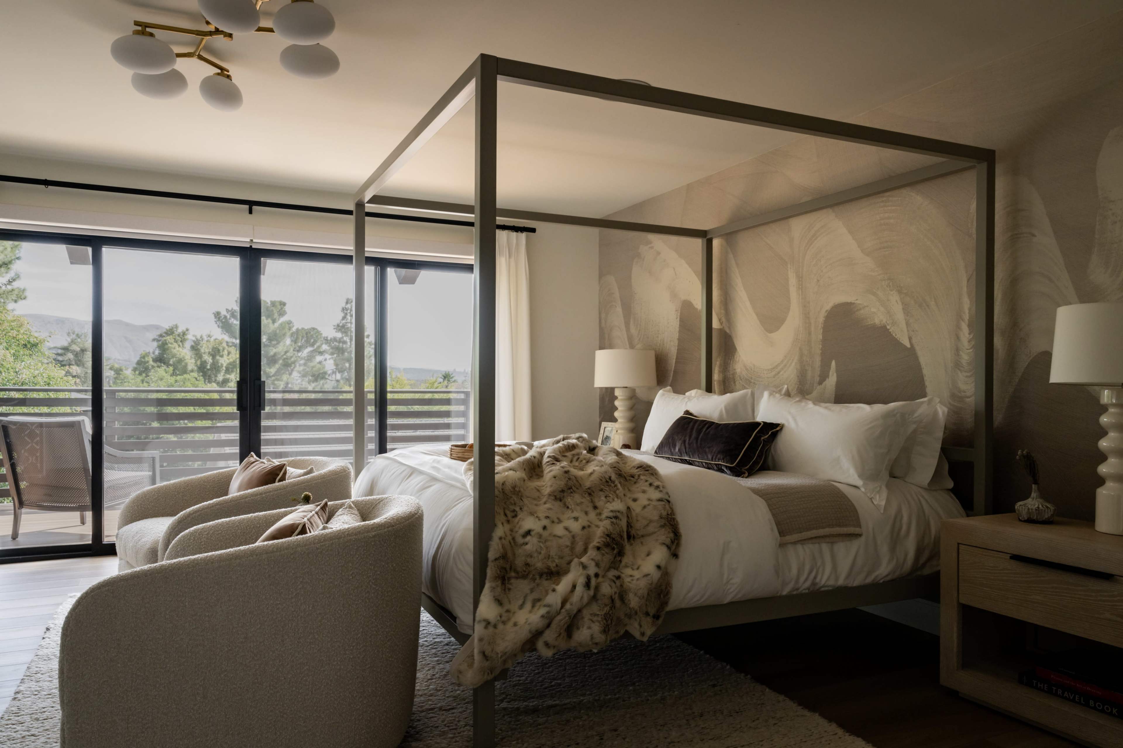 The image shows a modern bedroom with a four-poster bed, two upholstered chairs, and a balcony overlooking a green landscape.