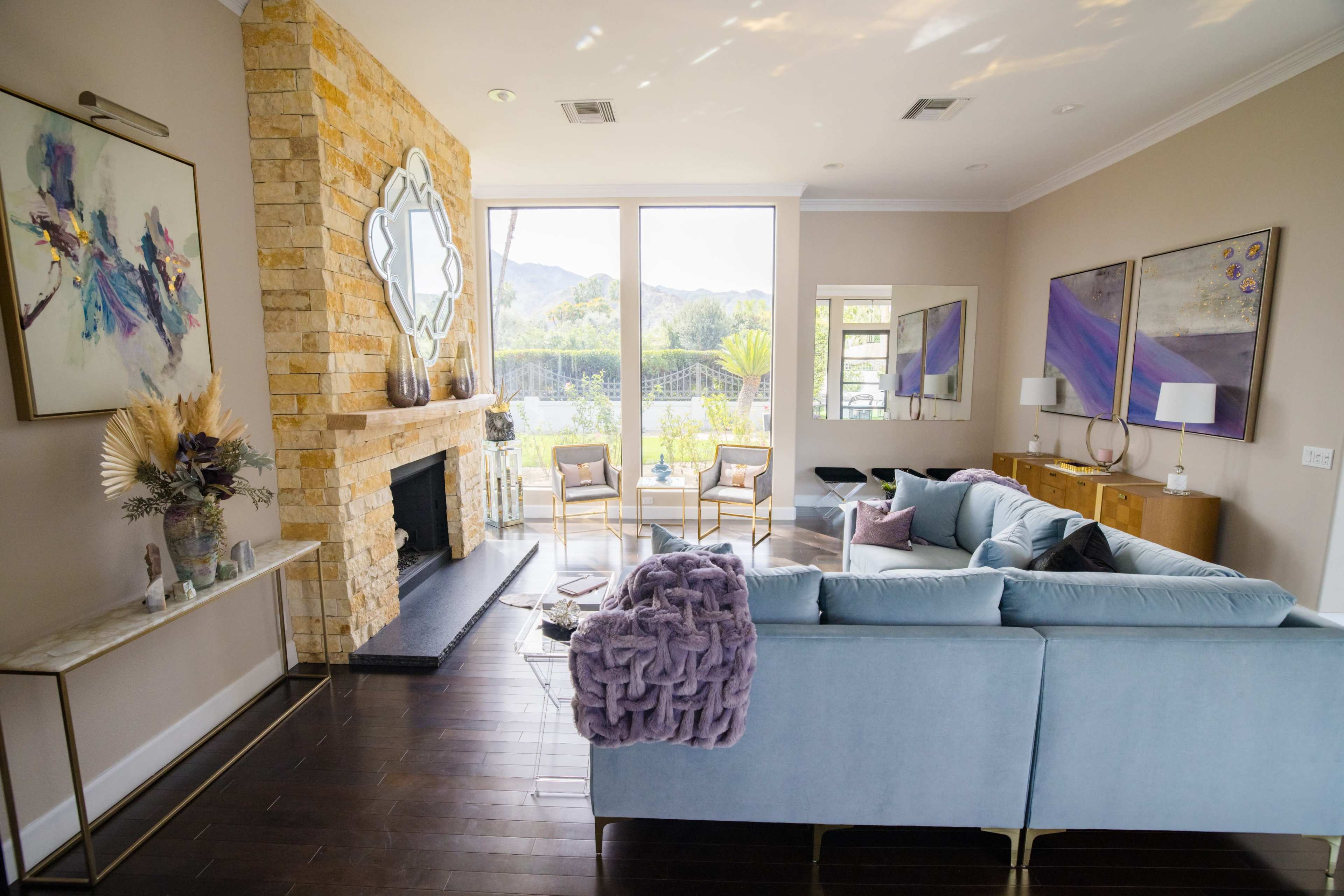 The room features a light-colored sofa with a textured throw, a stone fireplace, and large windows overlooking a patio and greenery.