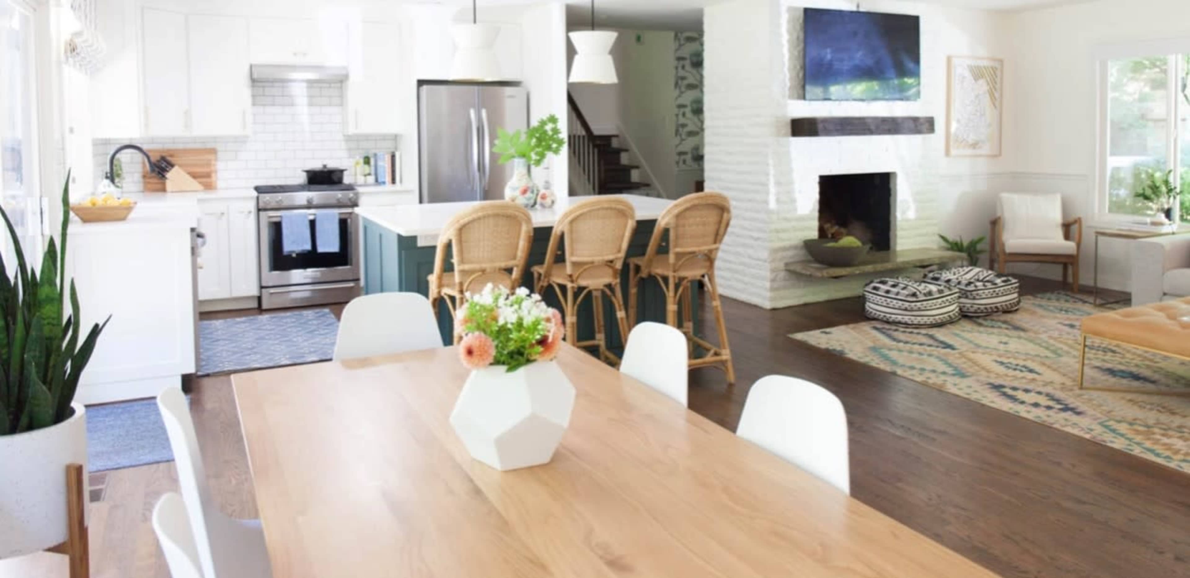 A modern open-concept kitchen and dining area featuring a wooden dining table, white cabinetry, a tiled backsplash, and a living space with a fireplace.