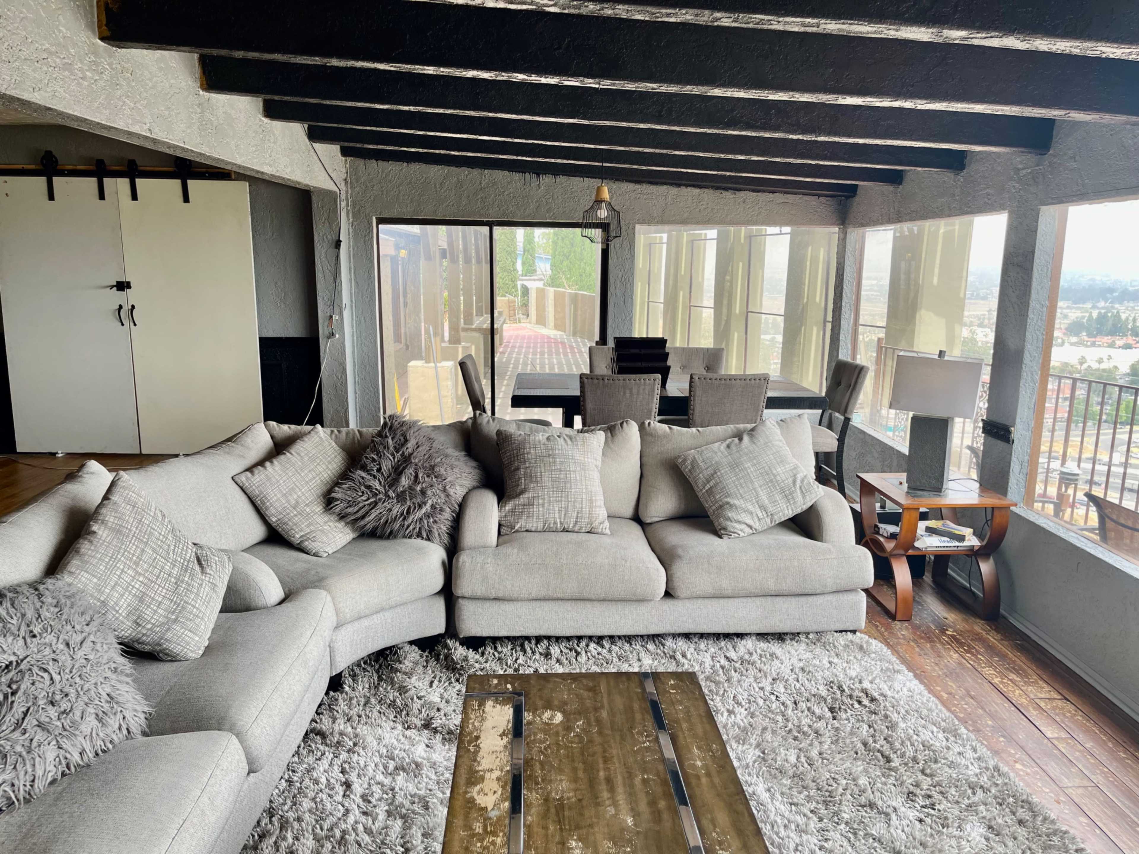 A light-colored sectional sofa with decorative pillows, a wooden coffee table, and large windows overlooking a scenic view, within a room that has exposed wooden beams and a cozy atmosphere.