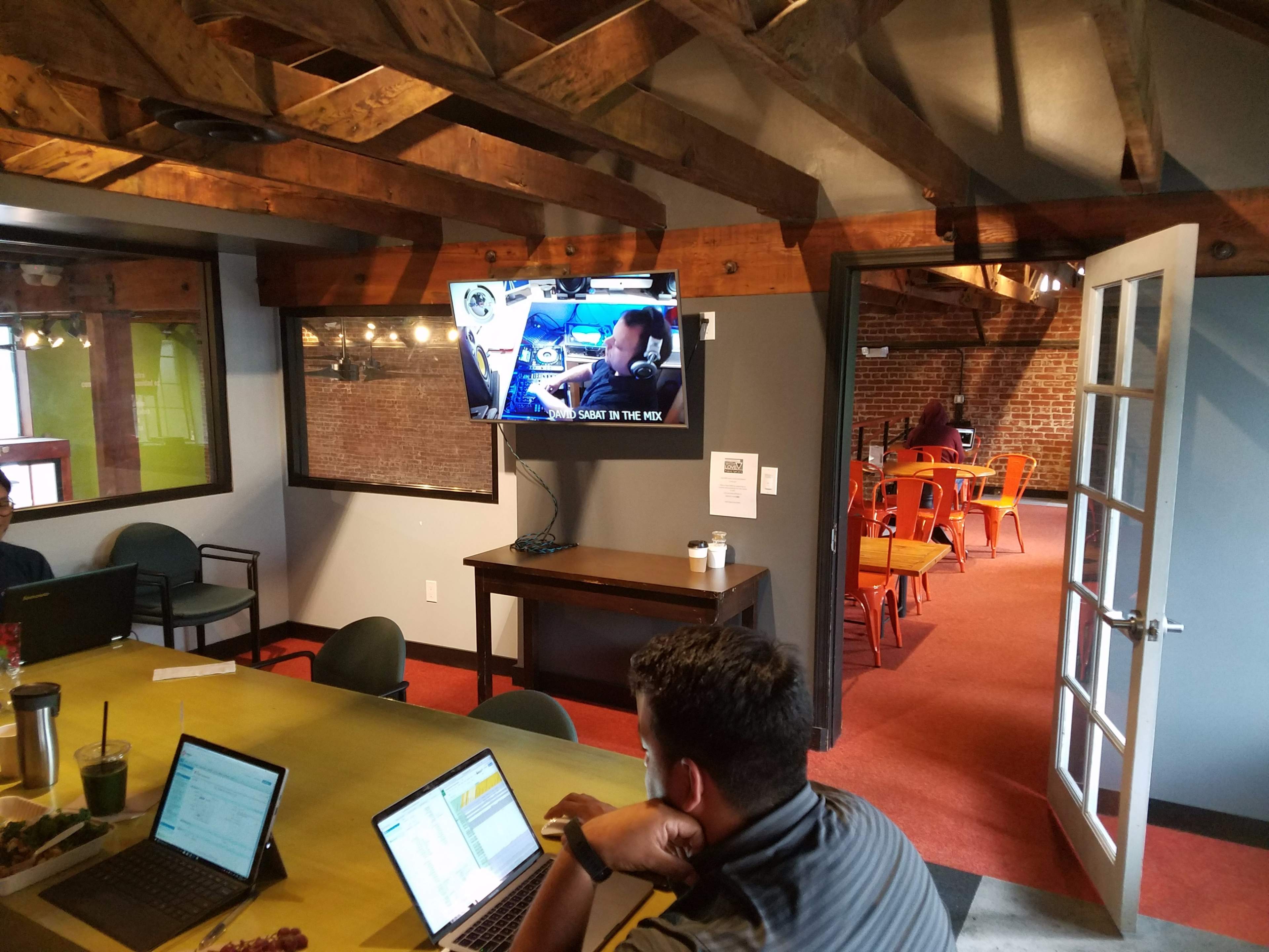 A person works on a laptop at a table while a television screen shows a video in a room with exposed wooden beams and brick walls.