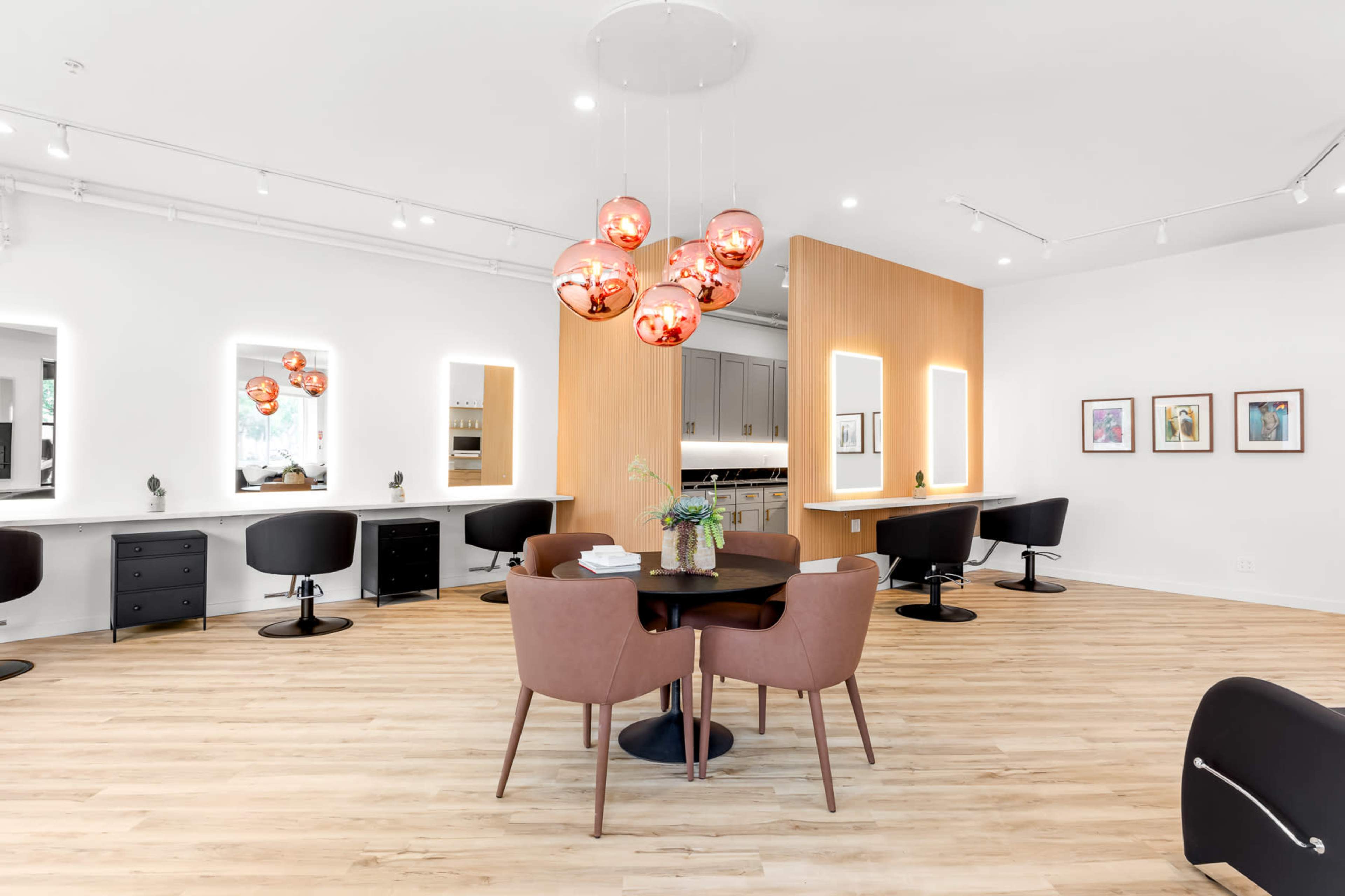 A bright hair salon features black styling chairs, a circular table with brown chairs, and decorative pendant lights hanging from the ceiling.