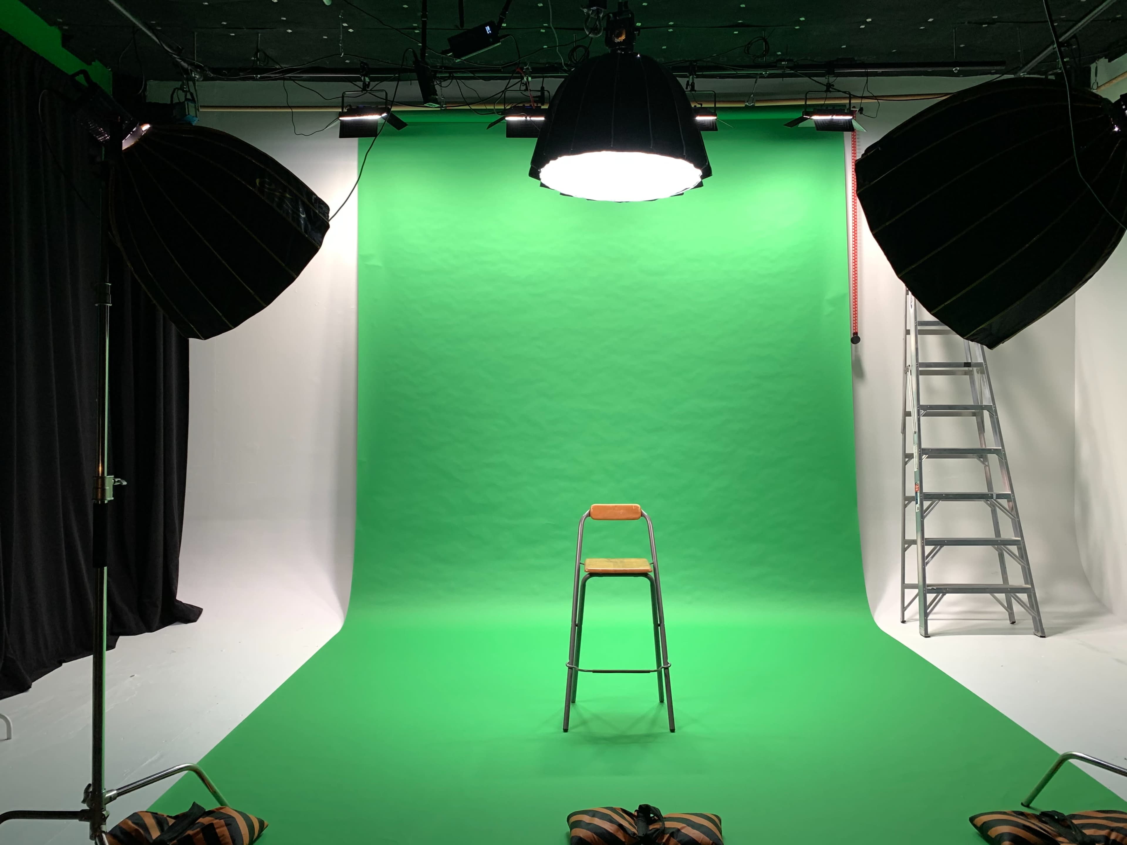 A studio setup features a green screen backdrop with a metal chair in the center, flanked by large studio lights and a ladder in the background.