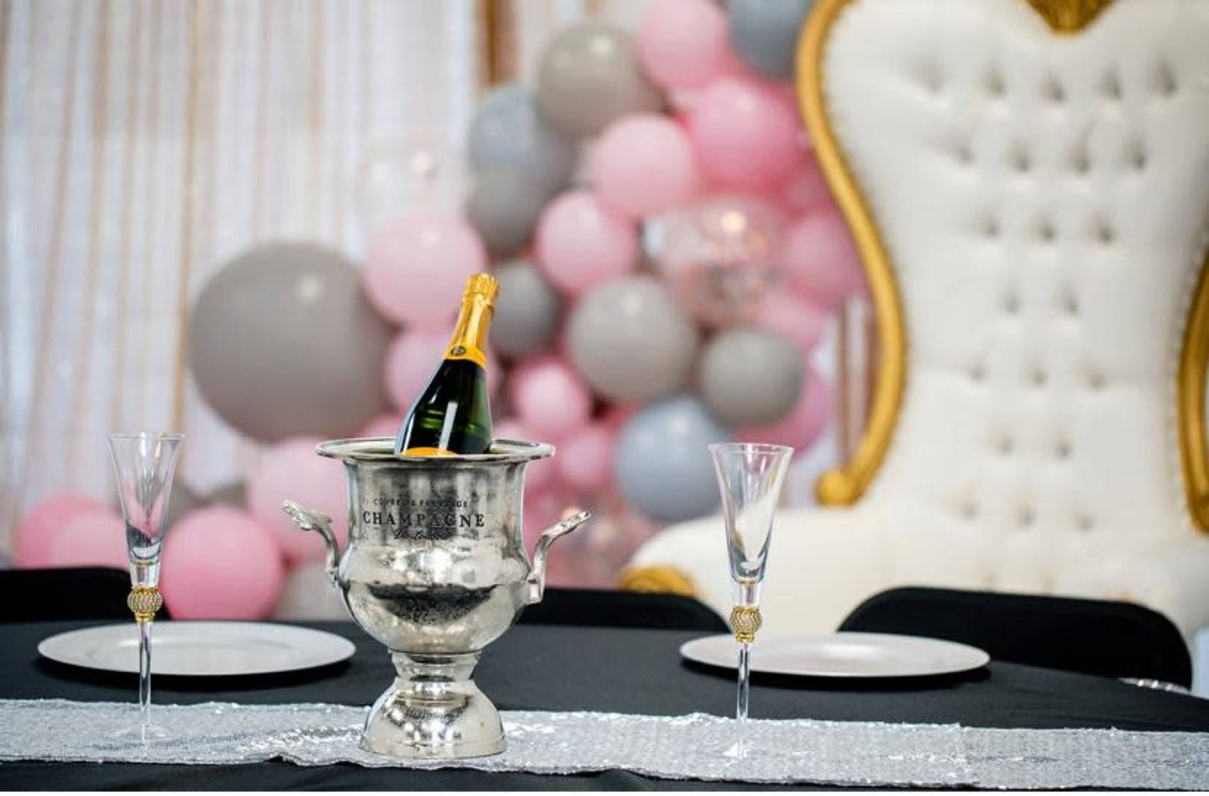 A silver champagne bucket with a bottle of champagne sits on a table with elegant glassware, while a backdrop of pink, gray, and white balloons adds decoration.