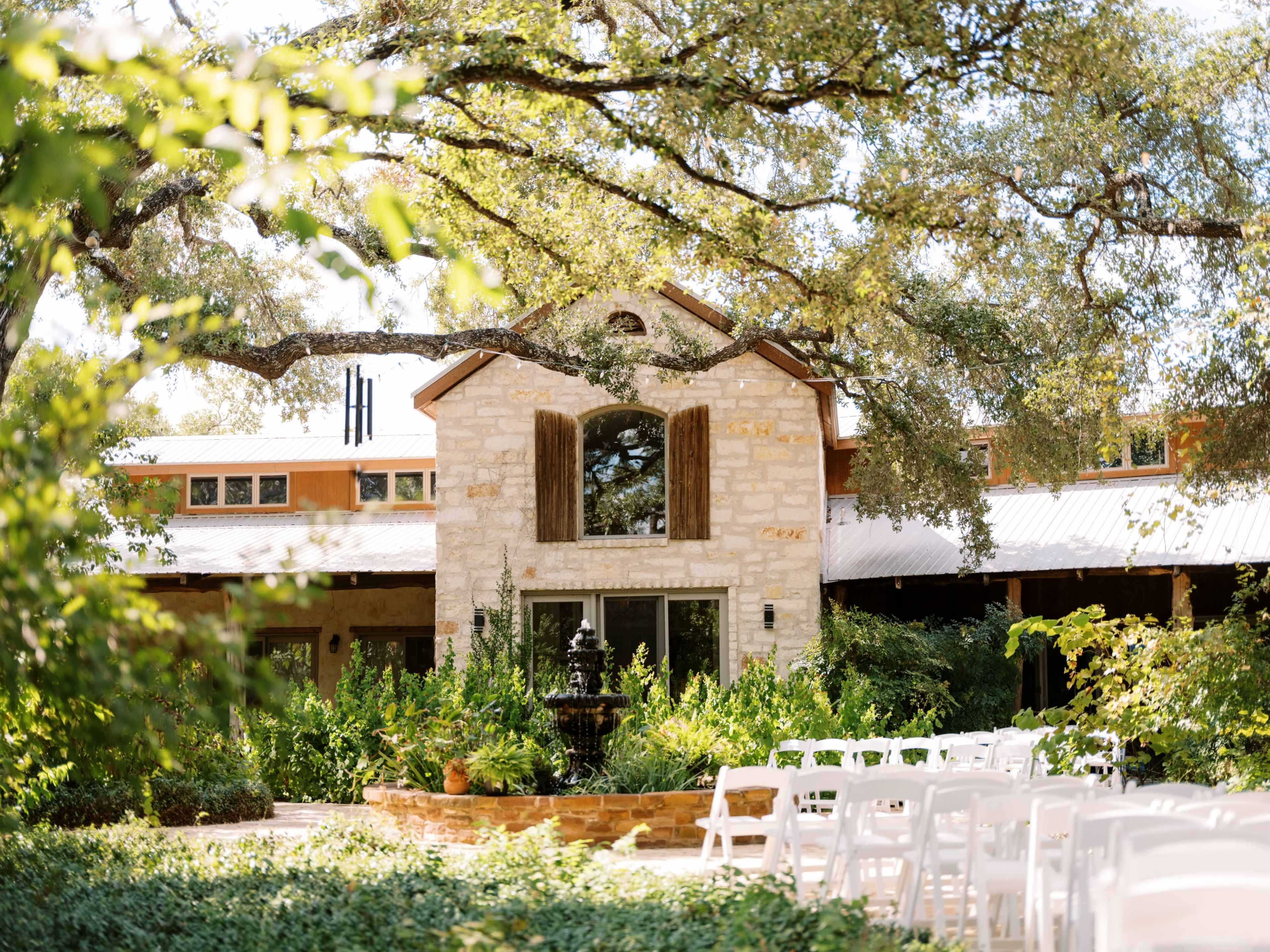 The image shows a landscaped outdoor space with white folding chairs arranged in front of a stone building featuring wooden shutters and a fountain surrounded by greenery.