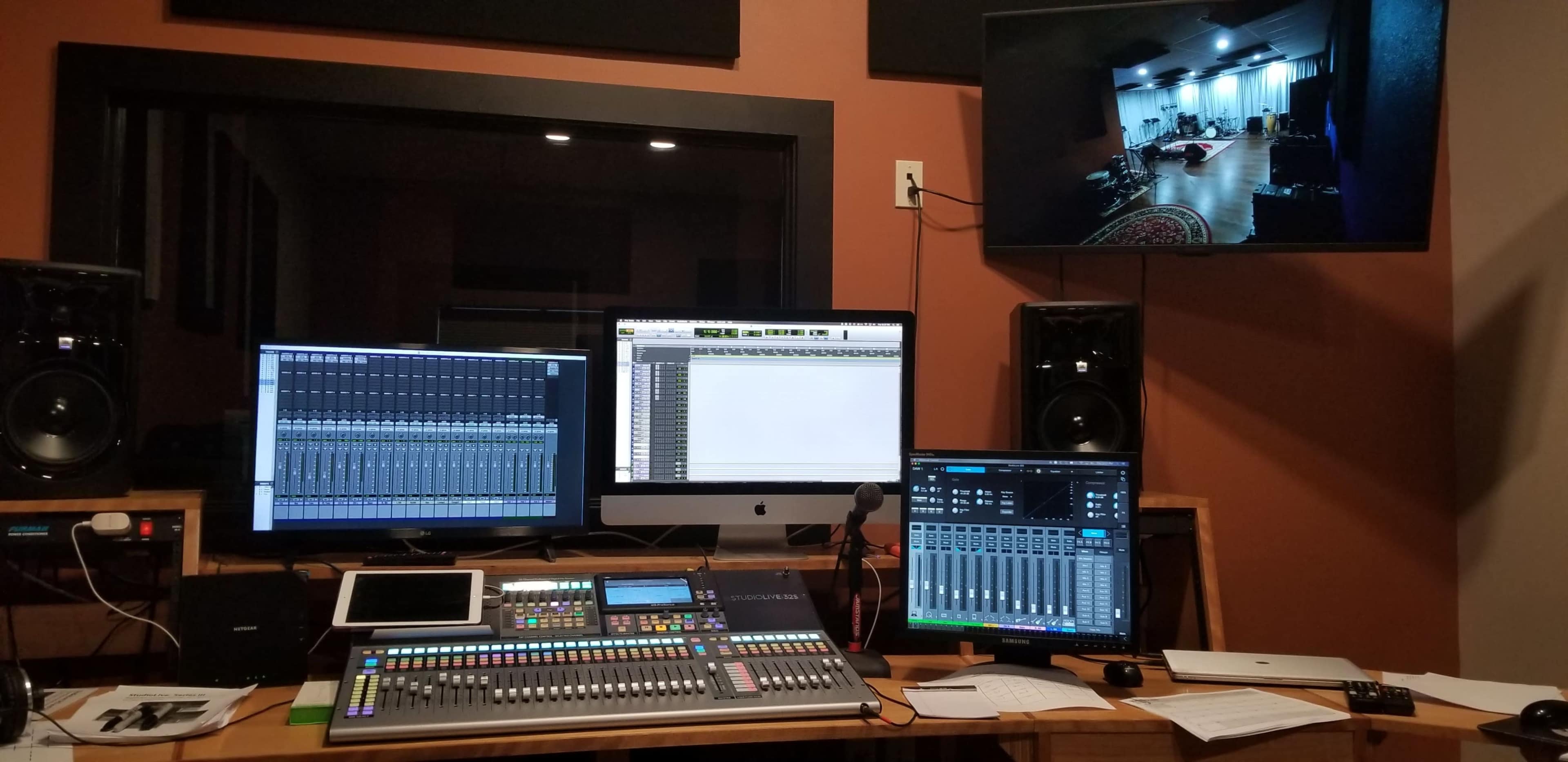 A music recording studio with multiple computer monitors, a digital mixing console, and a video feed of the studio space.