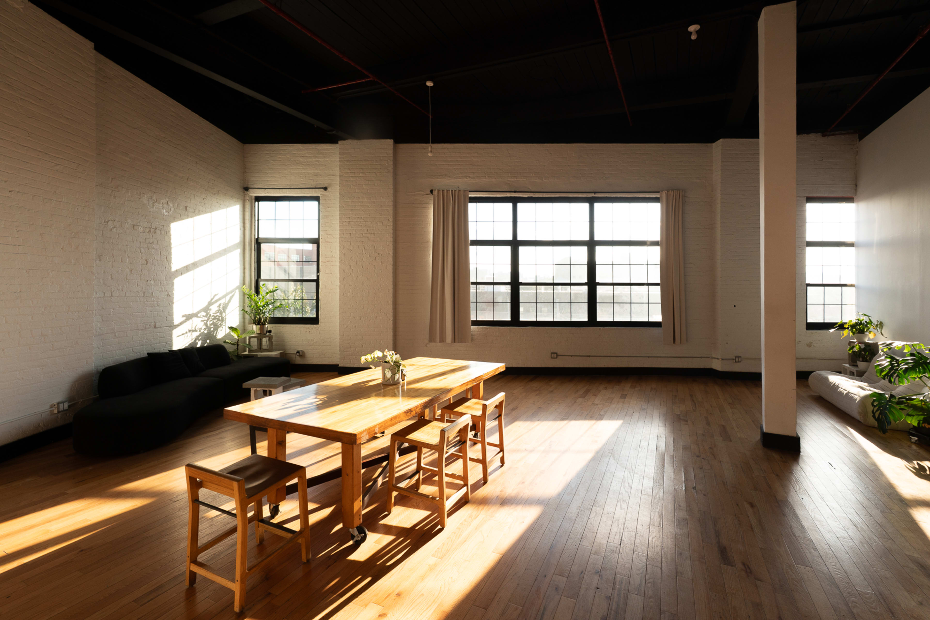 The image shows a spacious, bright room with large windows, a wooden dining table surrounded by stools, and a few plants in the corners.