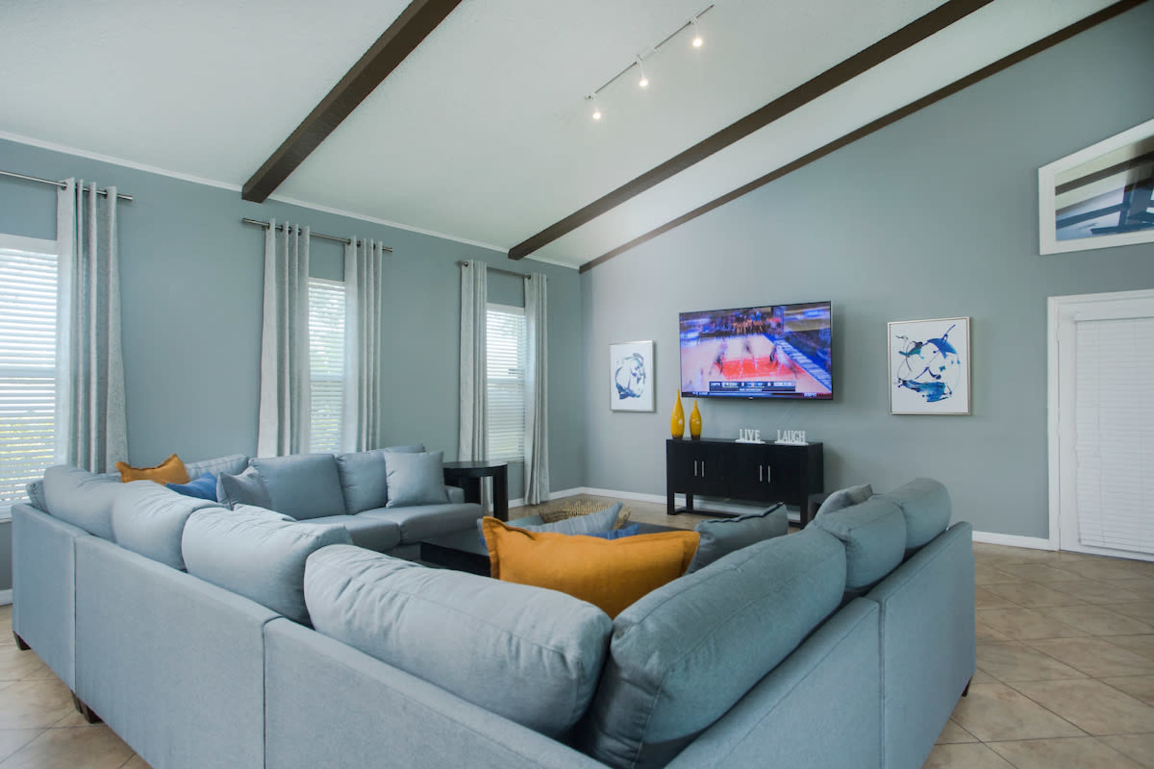 A spacious living room features a large sectional sofa, a wall-mounted television, and light gray walls with decorative art.