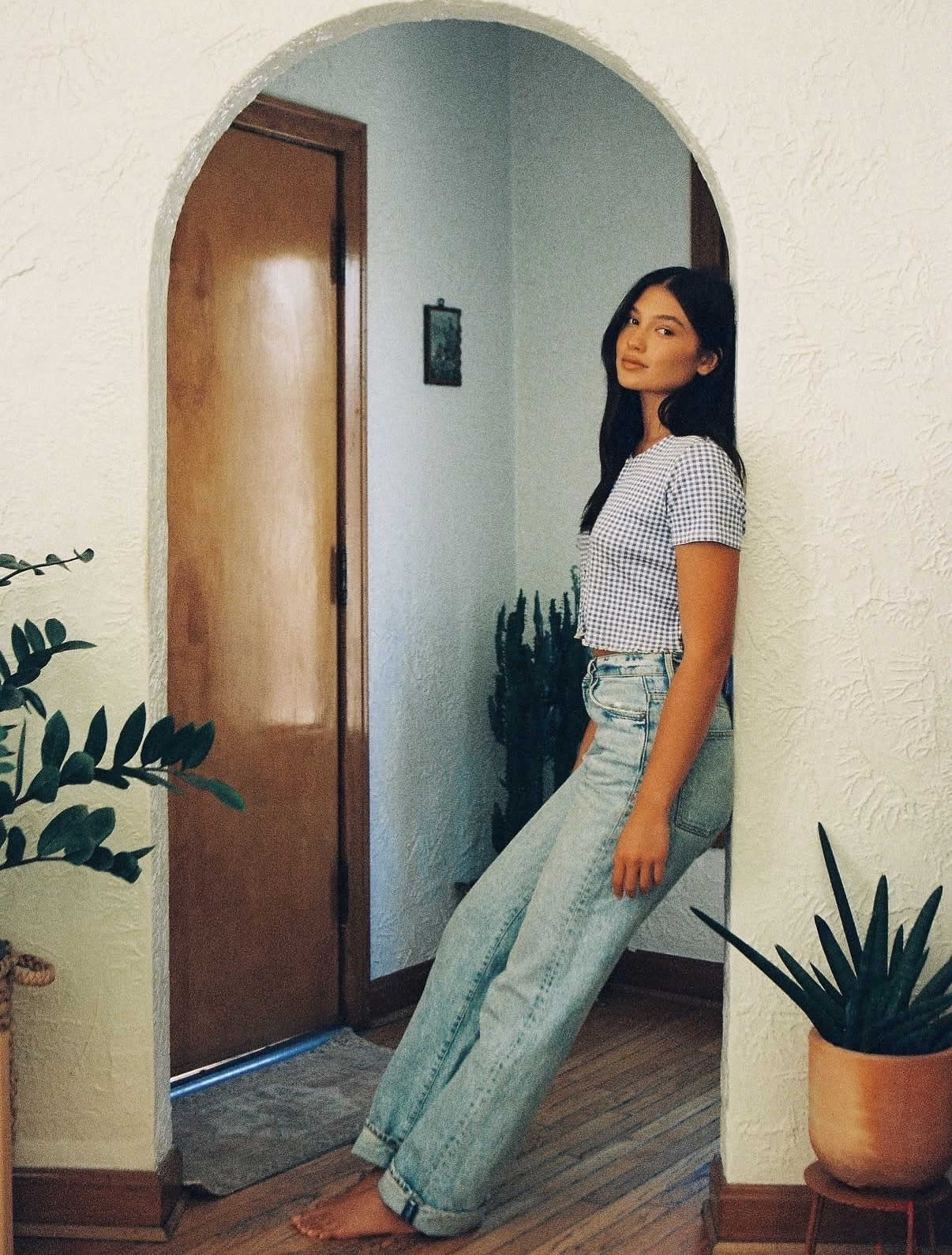 A person stands casually in a doorway with plants on either side of them, wearing a checkered shirt and high-waisted jeans.