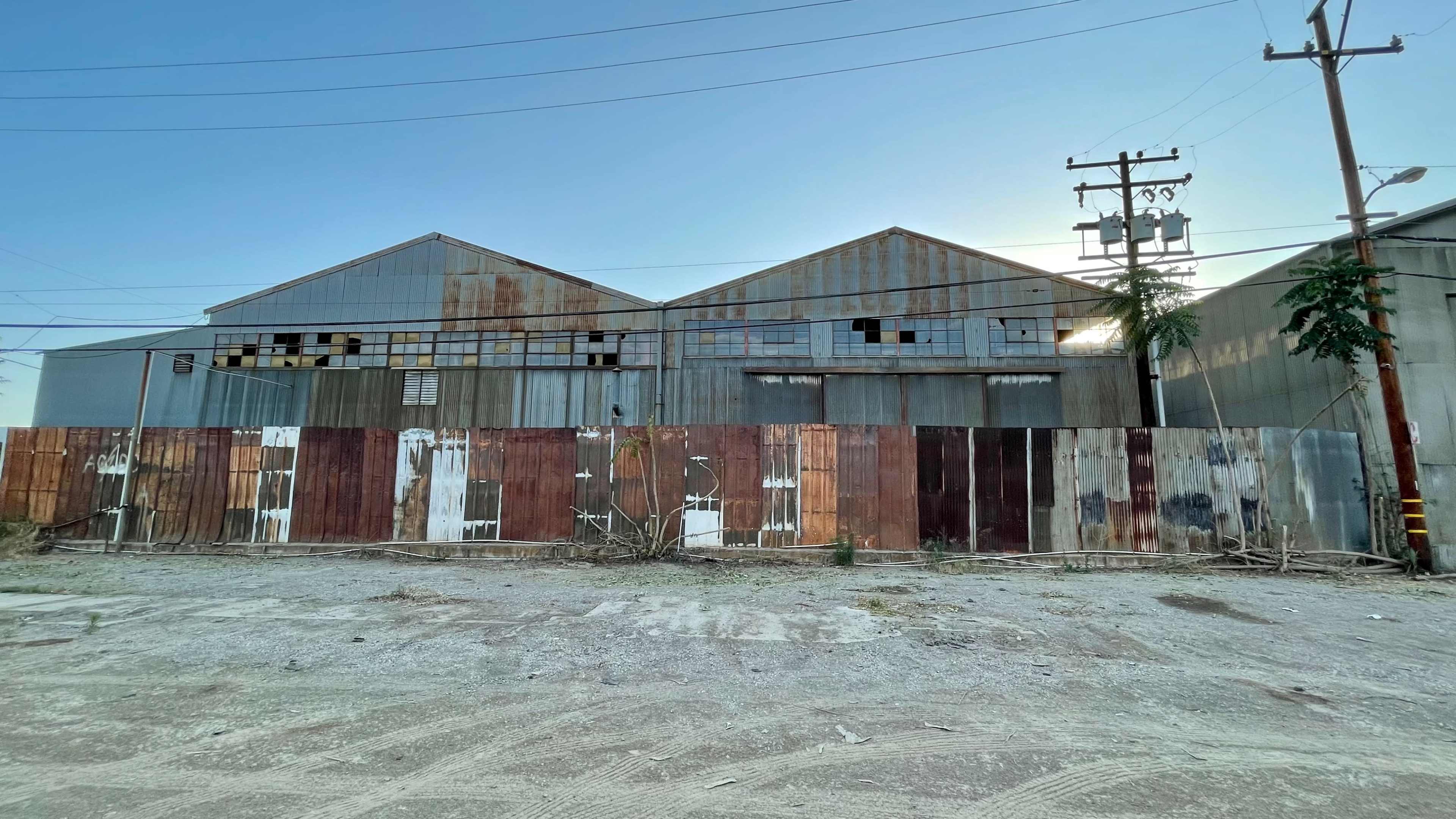 A pair of weathered industrial buildings behind a rusted metal fence in a gravel lot.