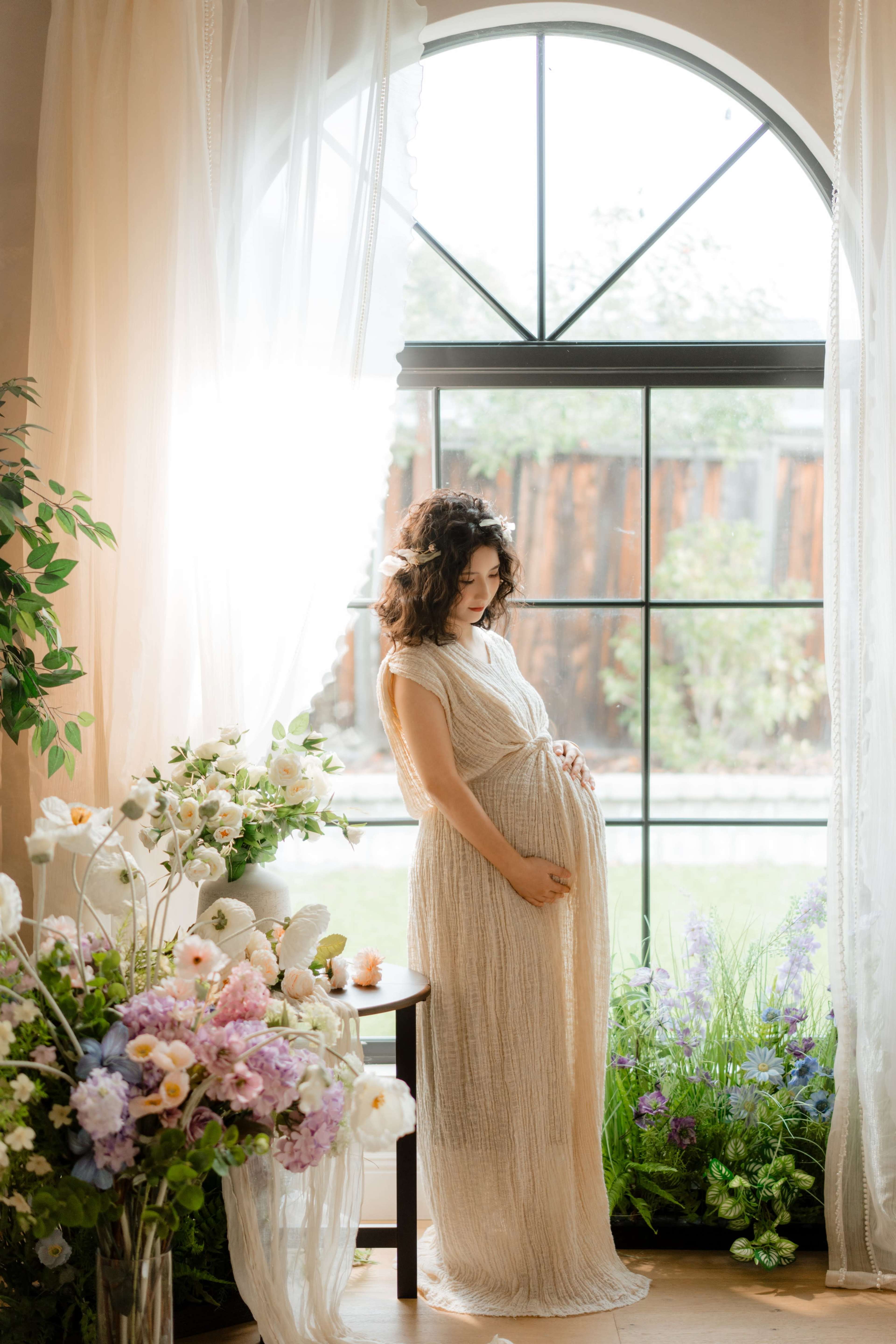 A pregnant woman in a flowing dress stands beside a table adorned with flowers, gazing out a large window with natural light streaming in.