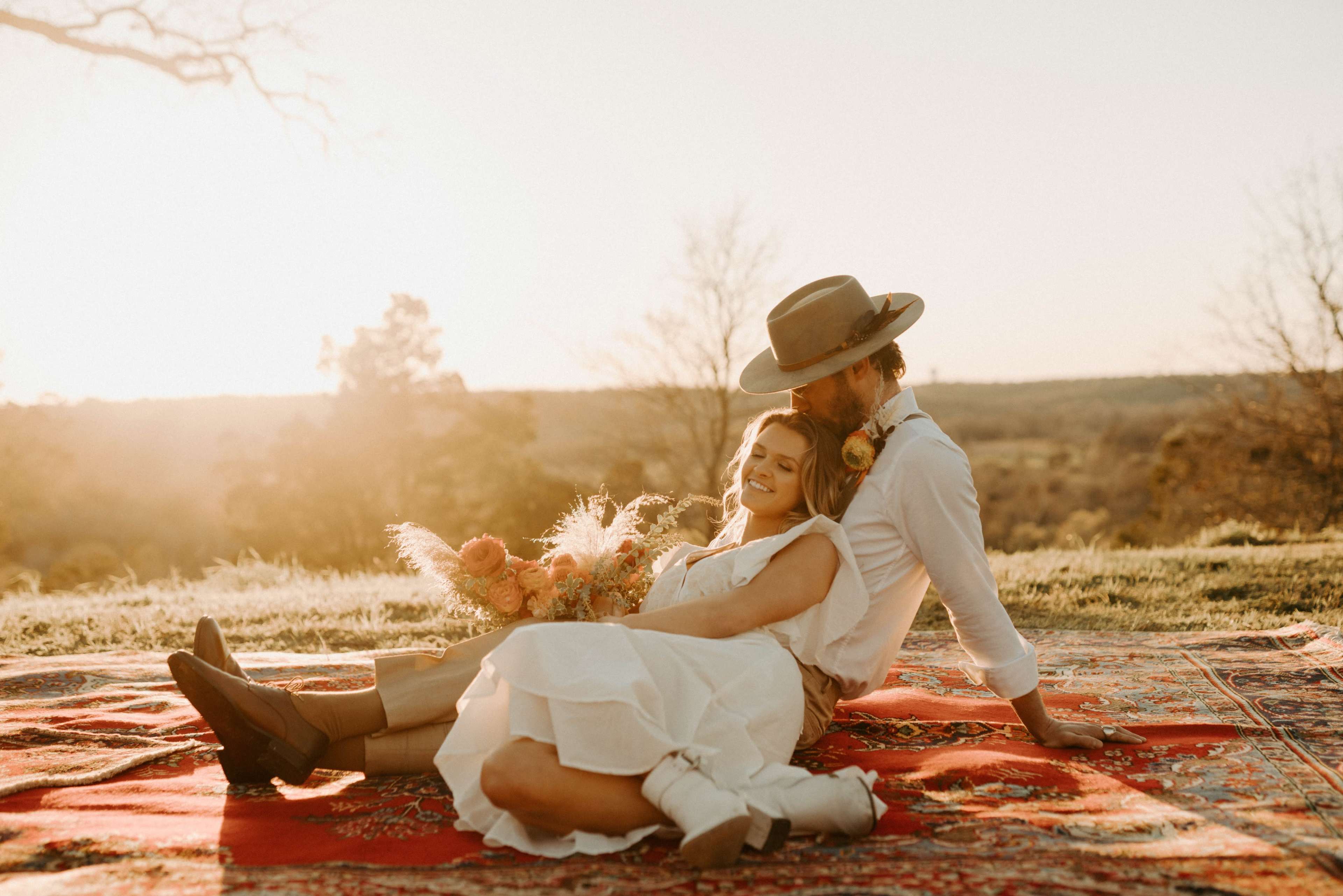 A couple sits on a patterned rug in a grassy landscape at sunset, with one person holding a bouquet of flowers.