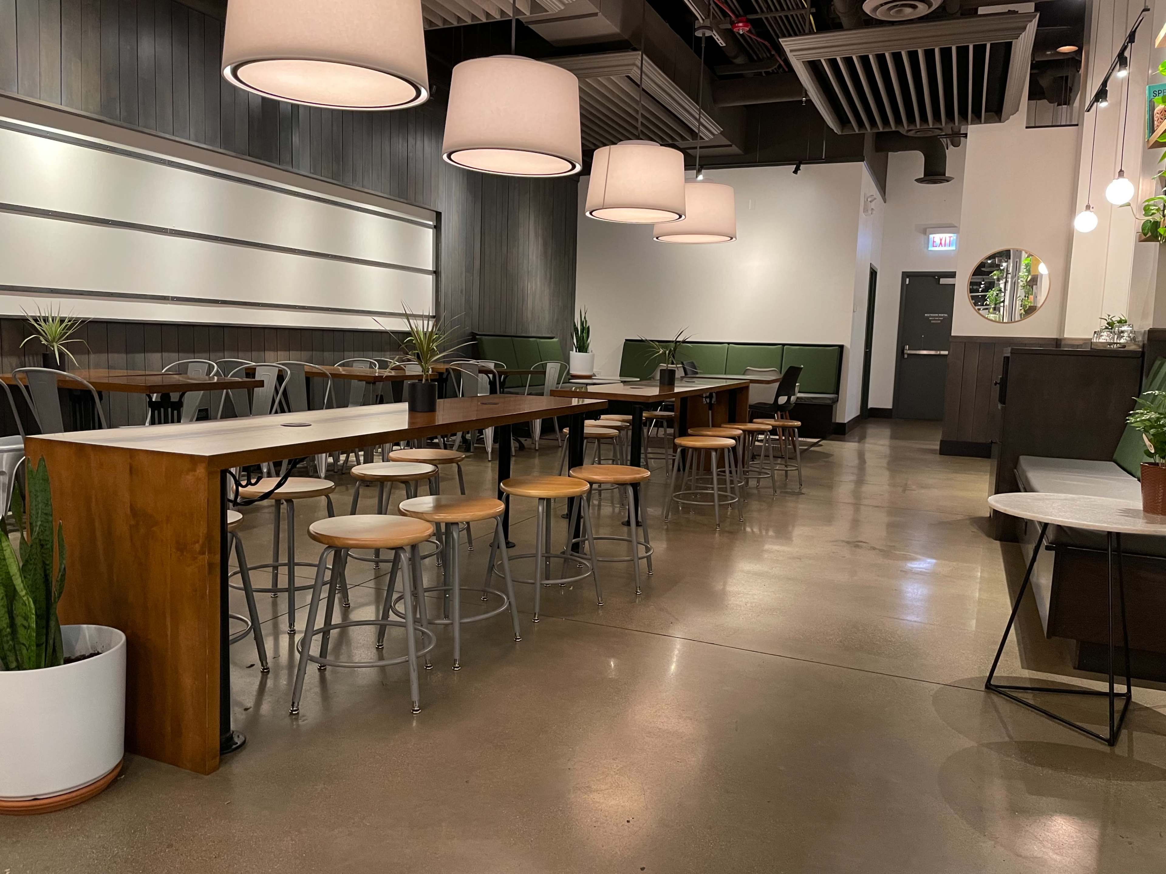 The image shows a modern café interior with wooden tables, metal chairs, and large pendant lights hanging from the ceiling.