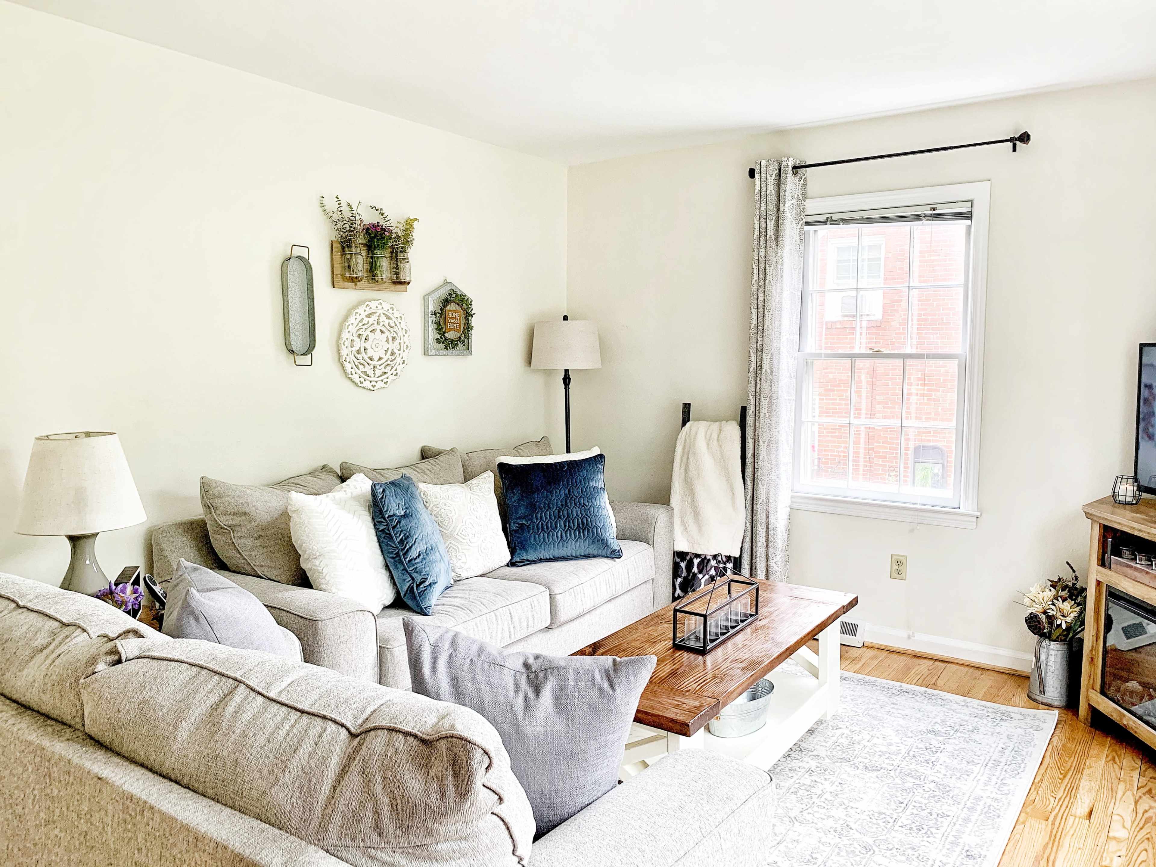 A cozy living room with light-colored sofas, a wooden coffee table, and decorative wall hangings near a window.