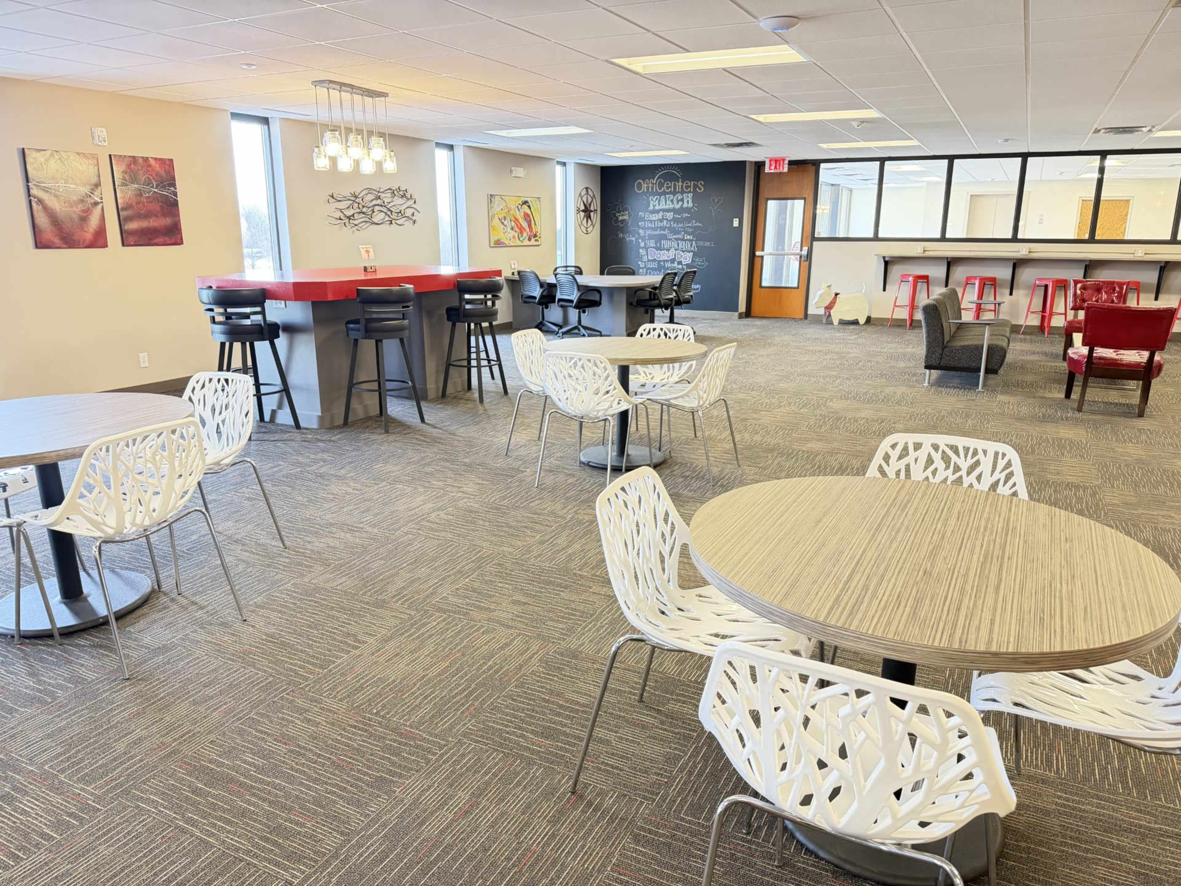 The image shows a spacious office lounge area featuring tables and chairs, a bar-style counter, and various seating arrangements.