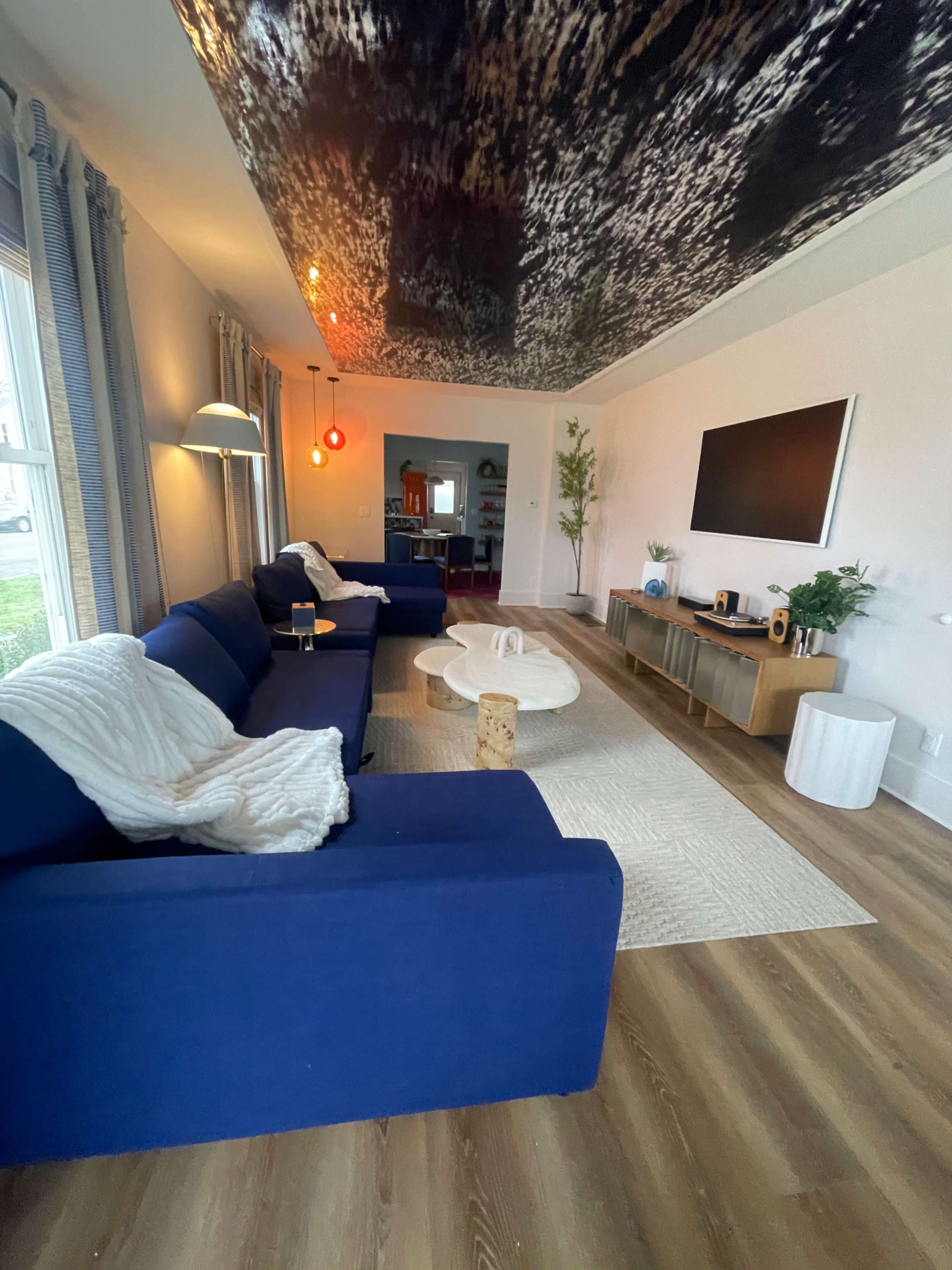 A living room with a blue sectional sofa, a circular coffee table, and a large television mounted on the wall, featuring a dark, textured ceiling.