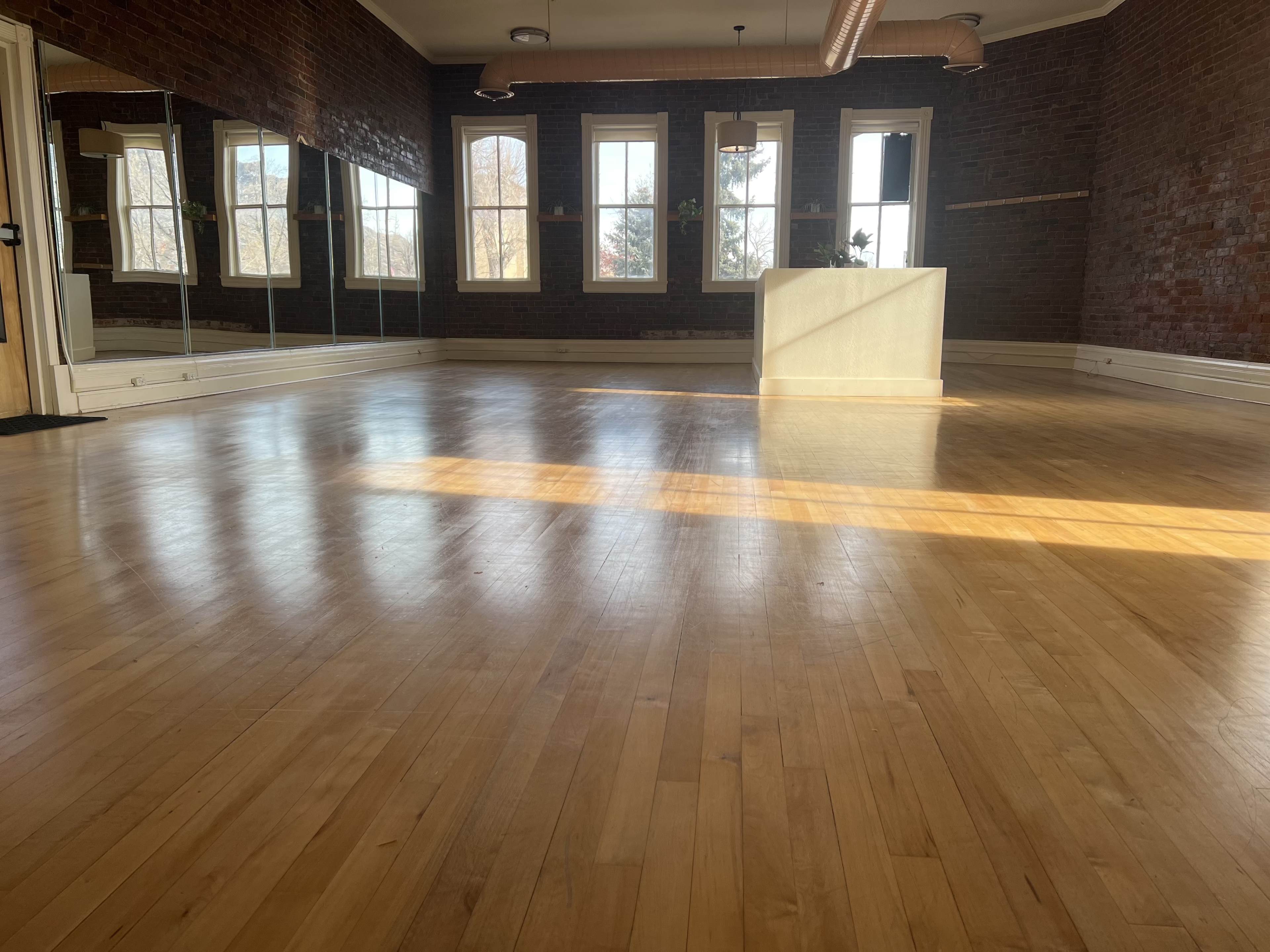 The image shows a spacious wooden-floored room with large windows and mirrored walls, illuminated by natural light.