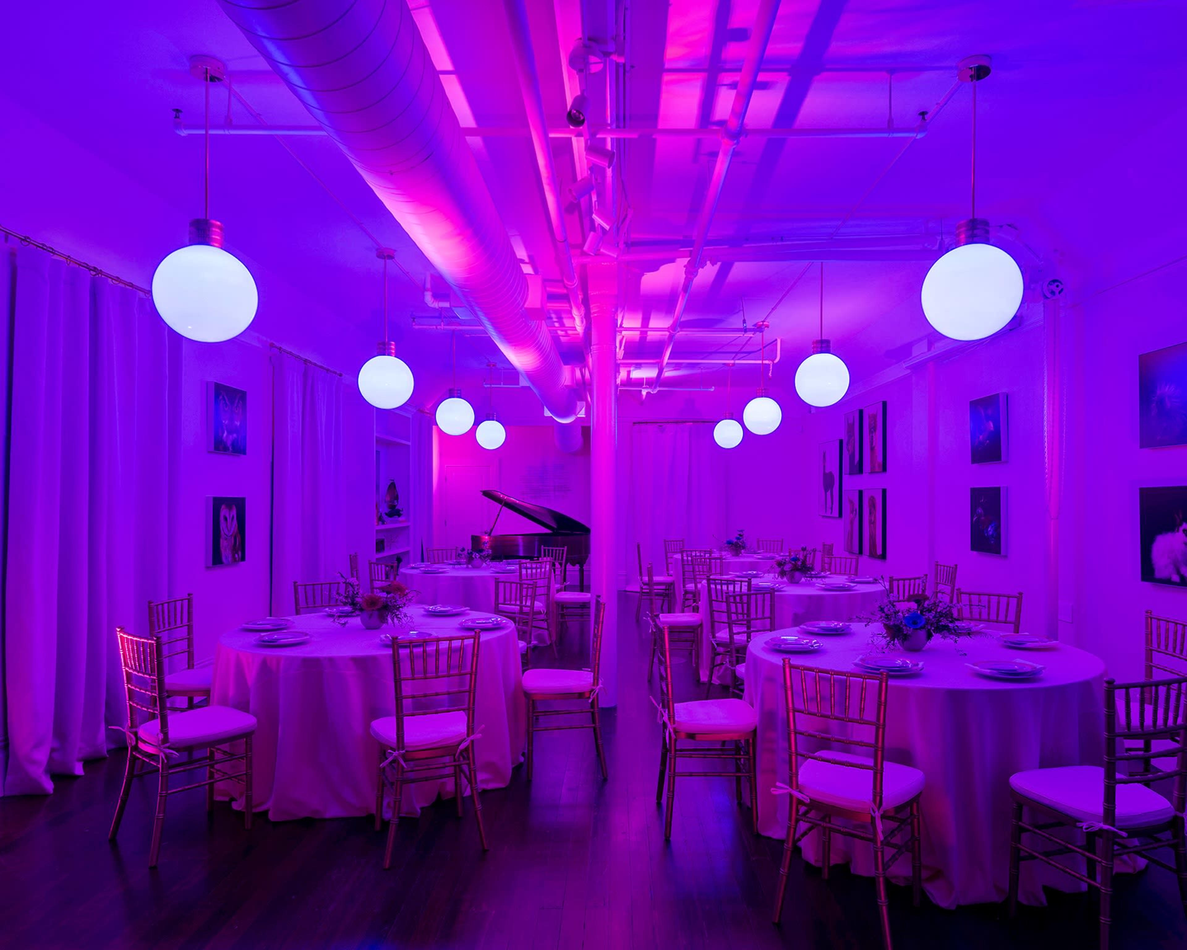 A decorated event space features circular tables with pink tablecloths, surrounded by gold chairs and illuminated by glowing purple lights.