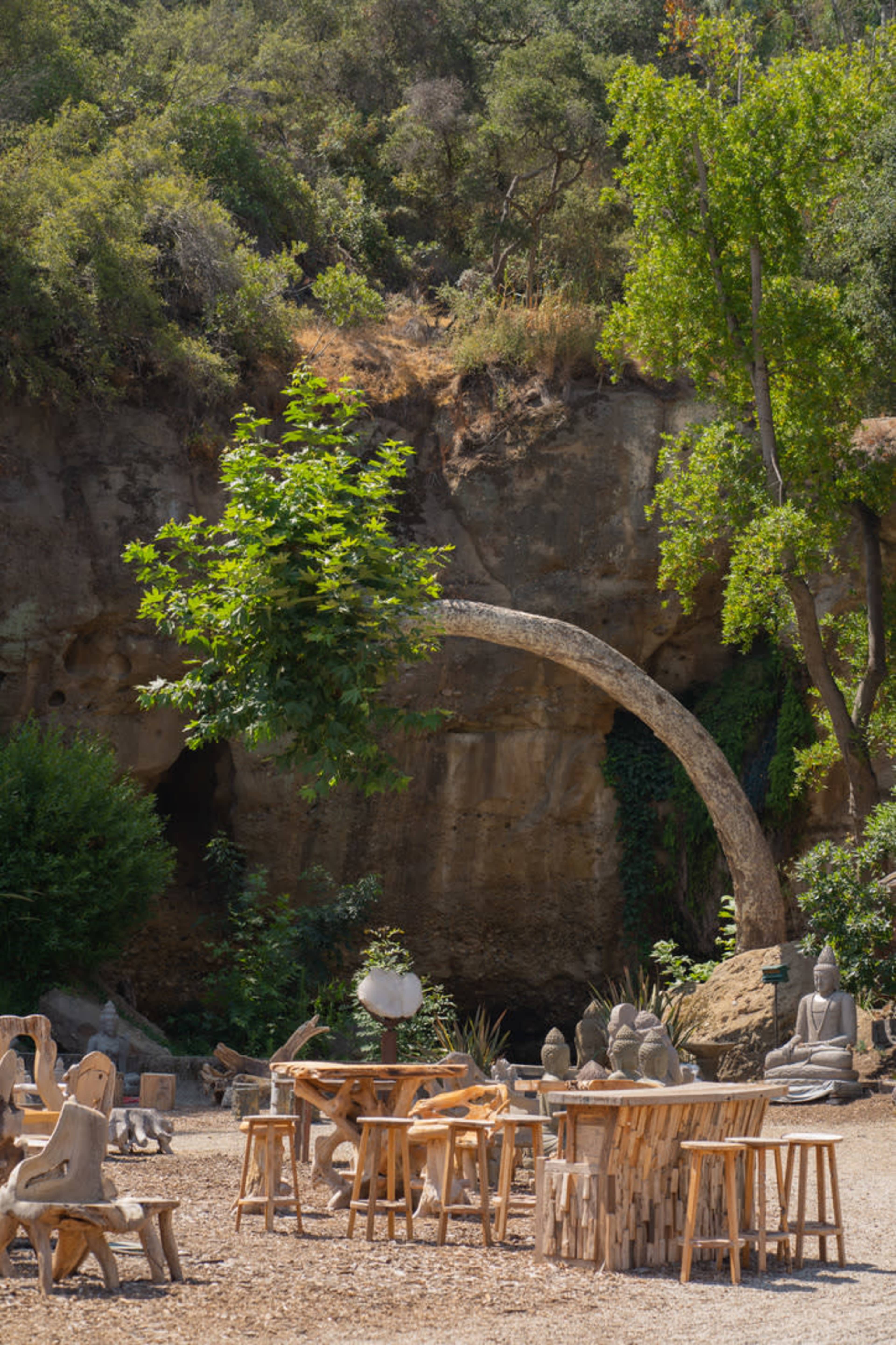 A serene outdoor setting features wooden tables and stools surrounded by sculpted figures and a hillside with trees.