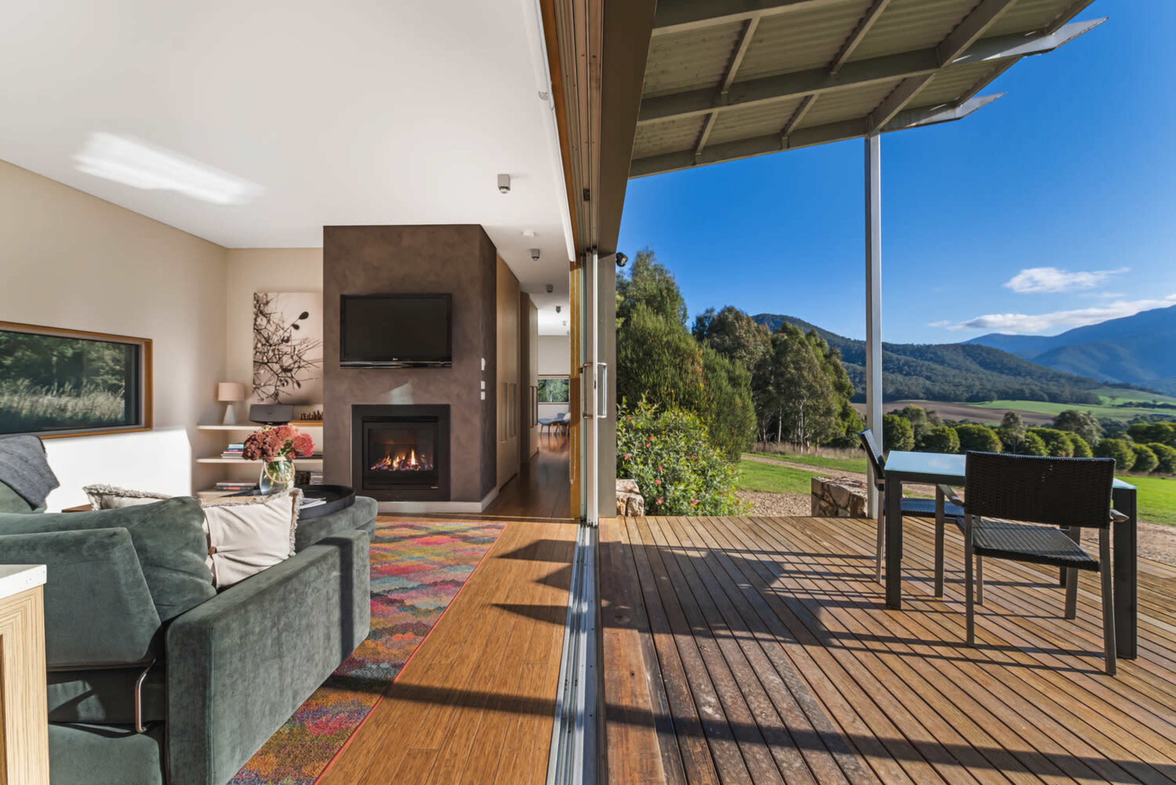 A modern living room with a sofa and a television, adjacent to a deck that overlooks a mountainous landscape.