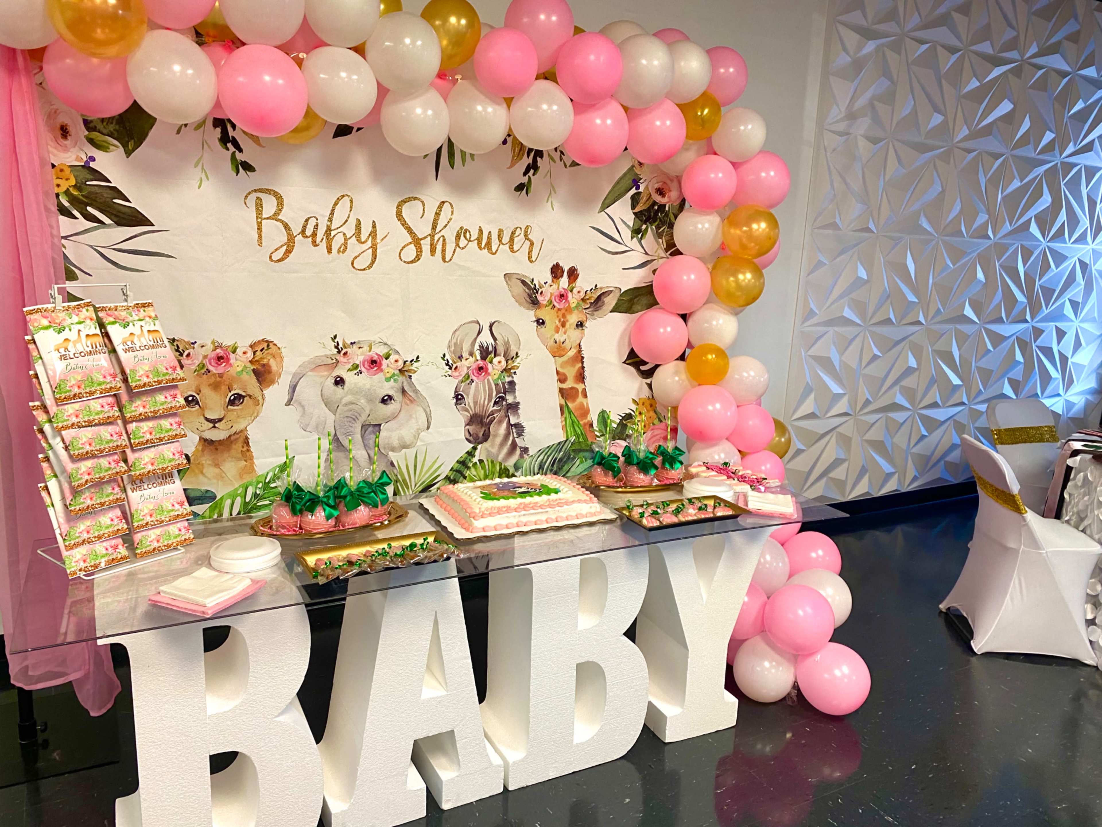 A decorated table for a baby shower featuring a cake, treats, and a backdrop with animal illustrations and balloons in pink and gold.