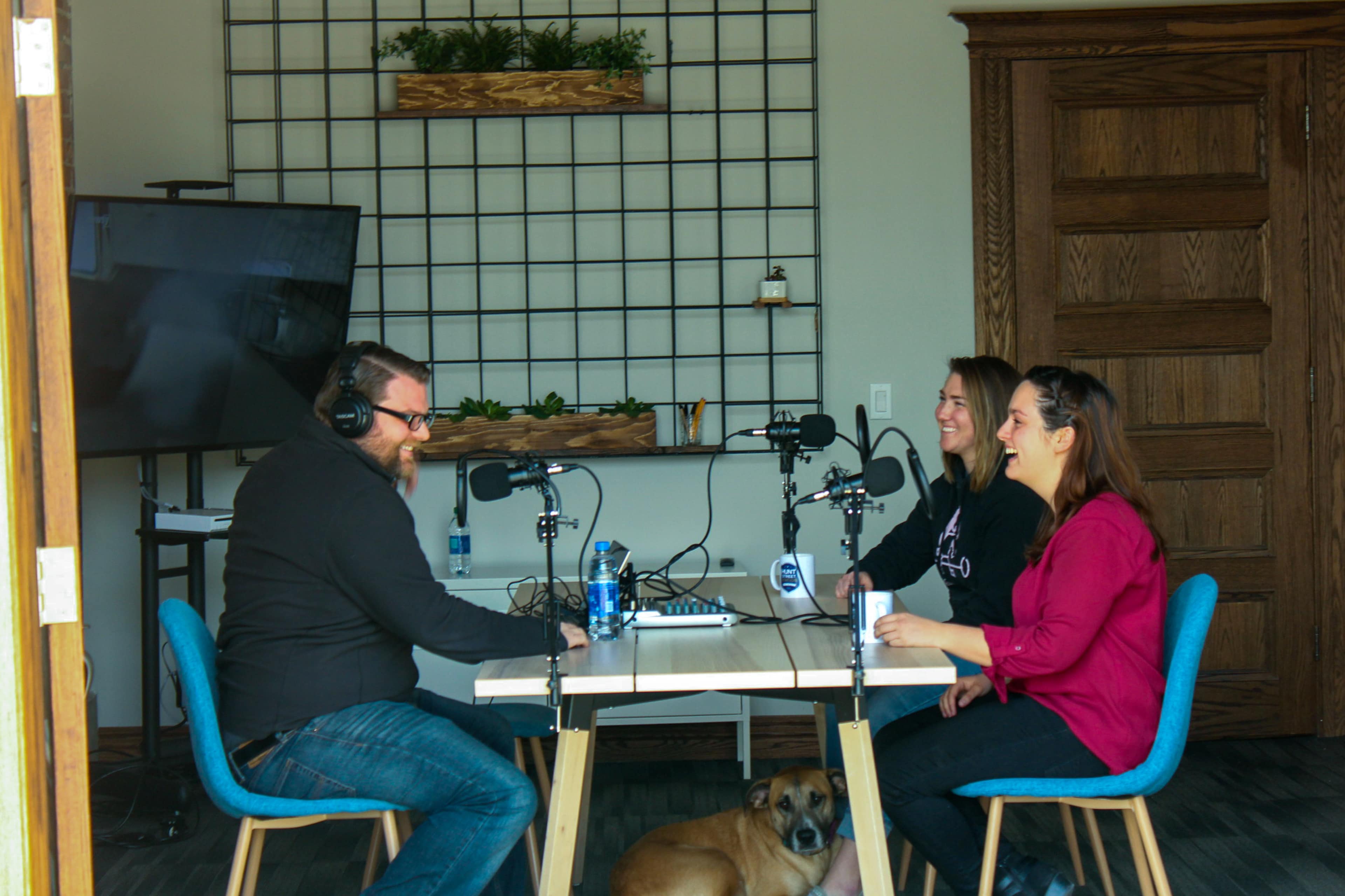 Three people sit at a table with microphones in a room featuring a grid wall and a large screen, while a dog lies on the floor.