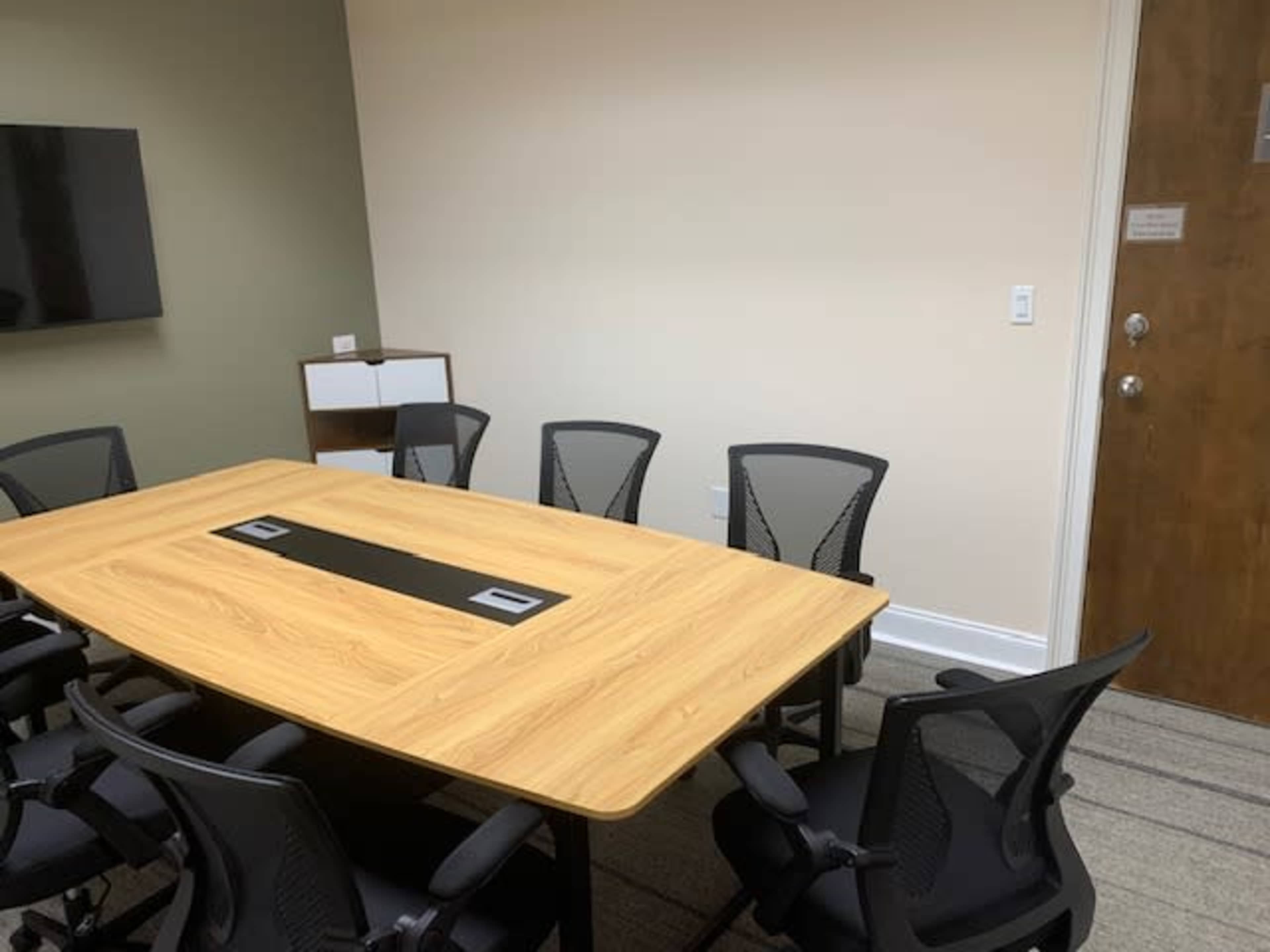 A large wooden conference table surrounded by black mesh chairs is situated in a brightly lit meeting room with a TV on the wall and a door visible in the background.