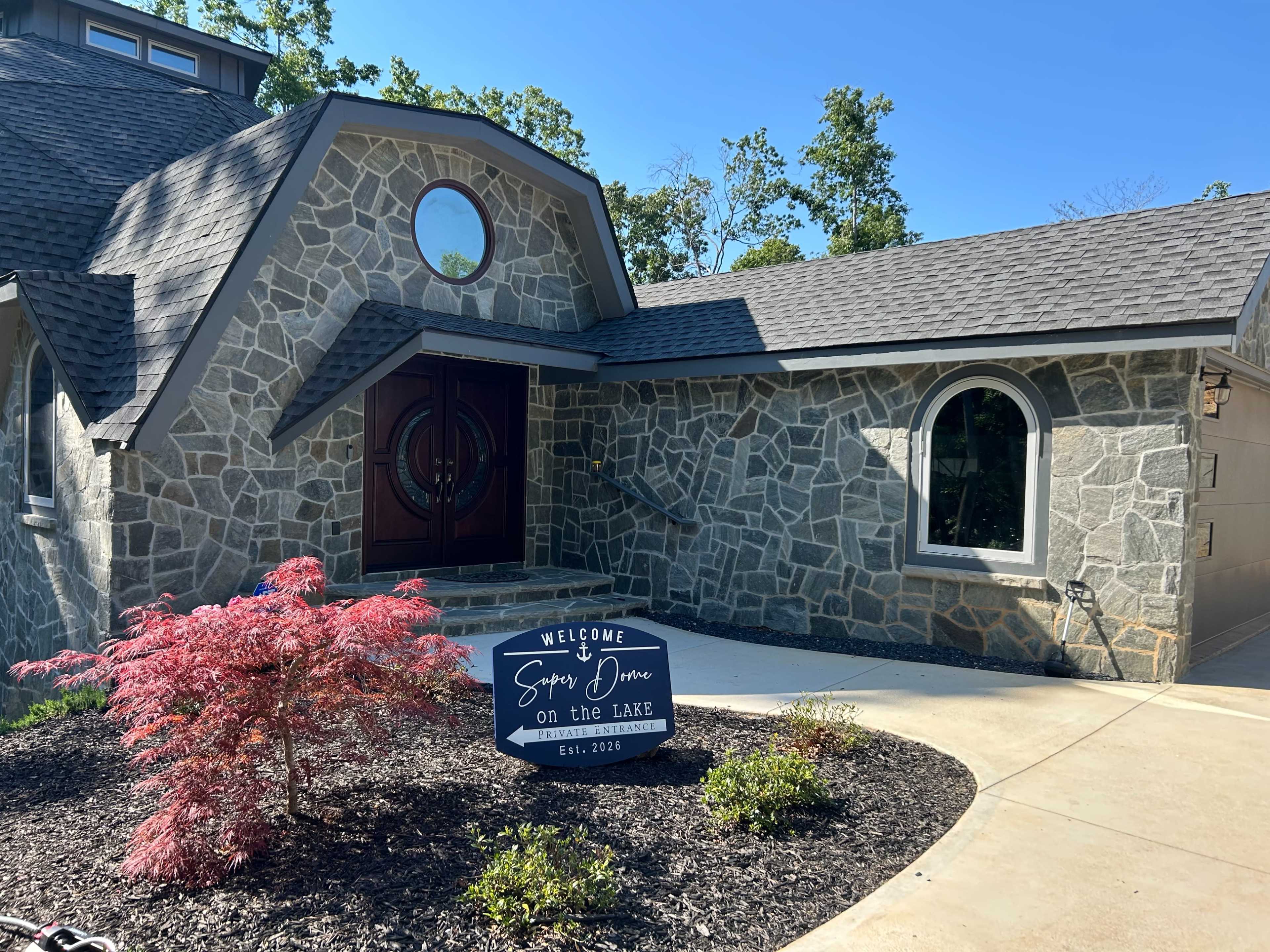 The image shows a stone-faced house with a circular window and a welcoming sign in front, surrounded by landscaped greenery.