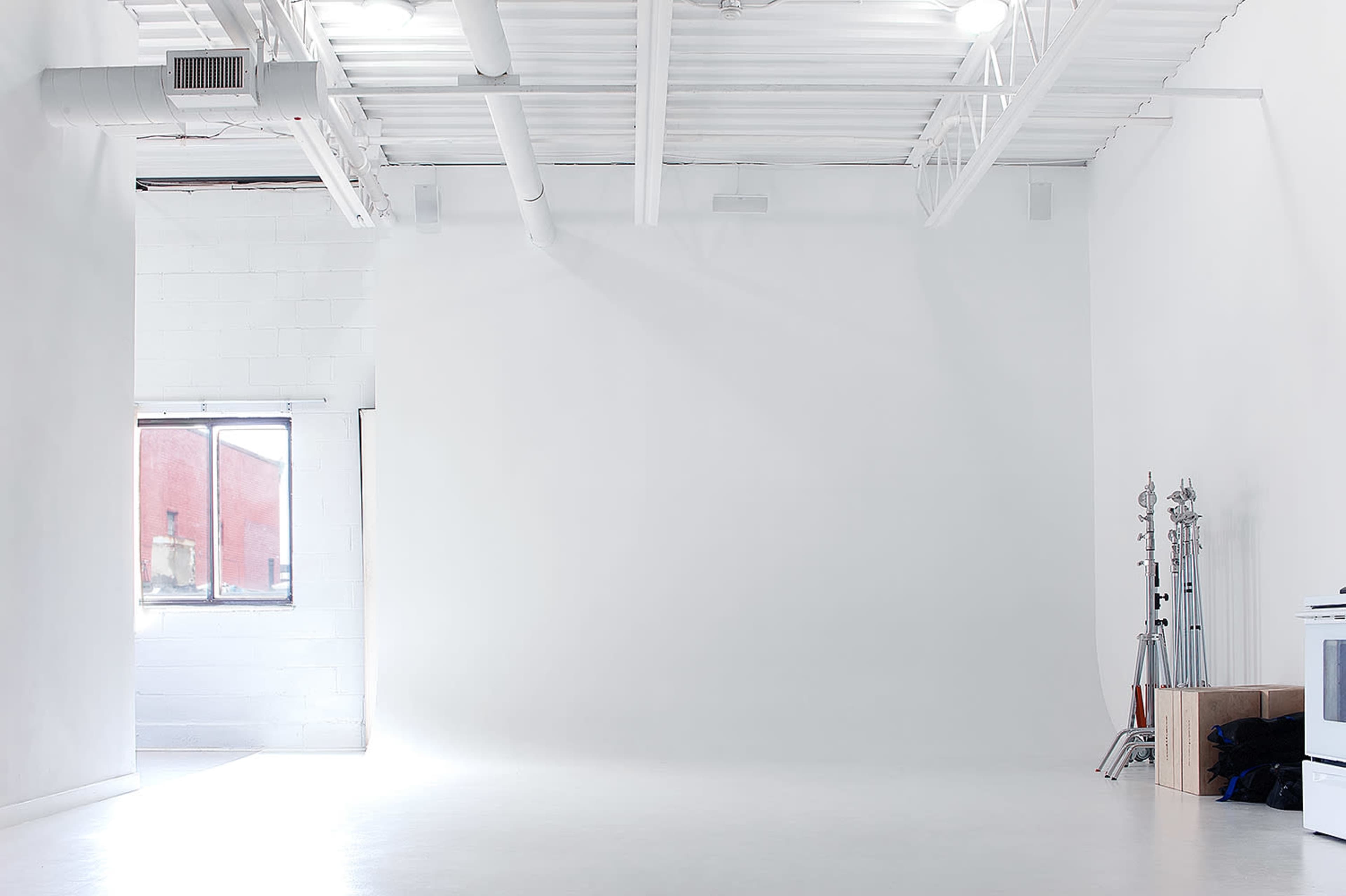 The image shows a spacious, well-lit photography studio with white walls, a window on the left, and equipment stands on the right.