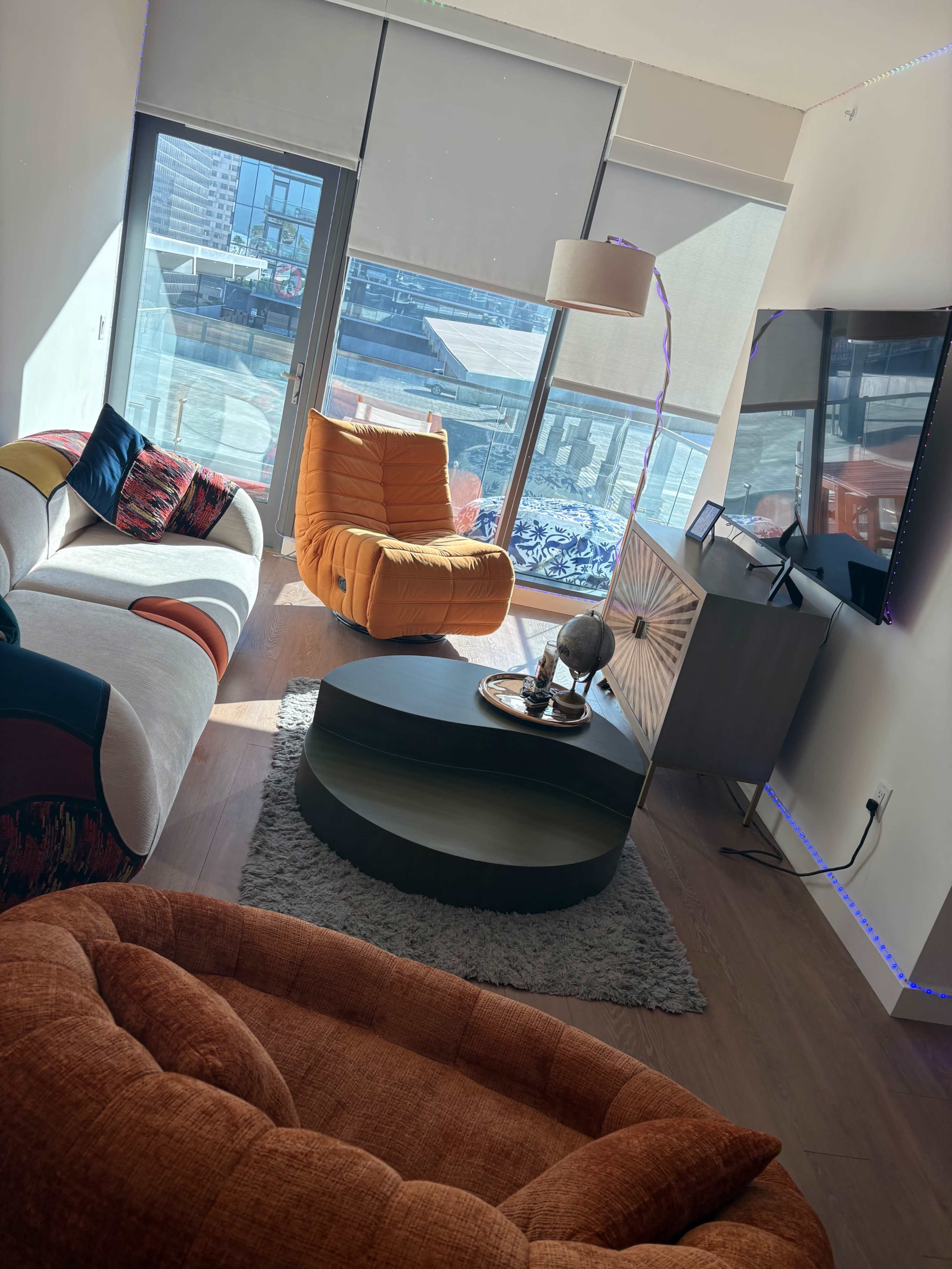 The image shows a modern living room with colorful furniture, including a circular coffee table and large windows letting in natural light.