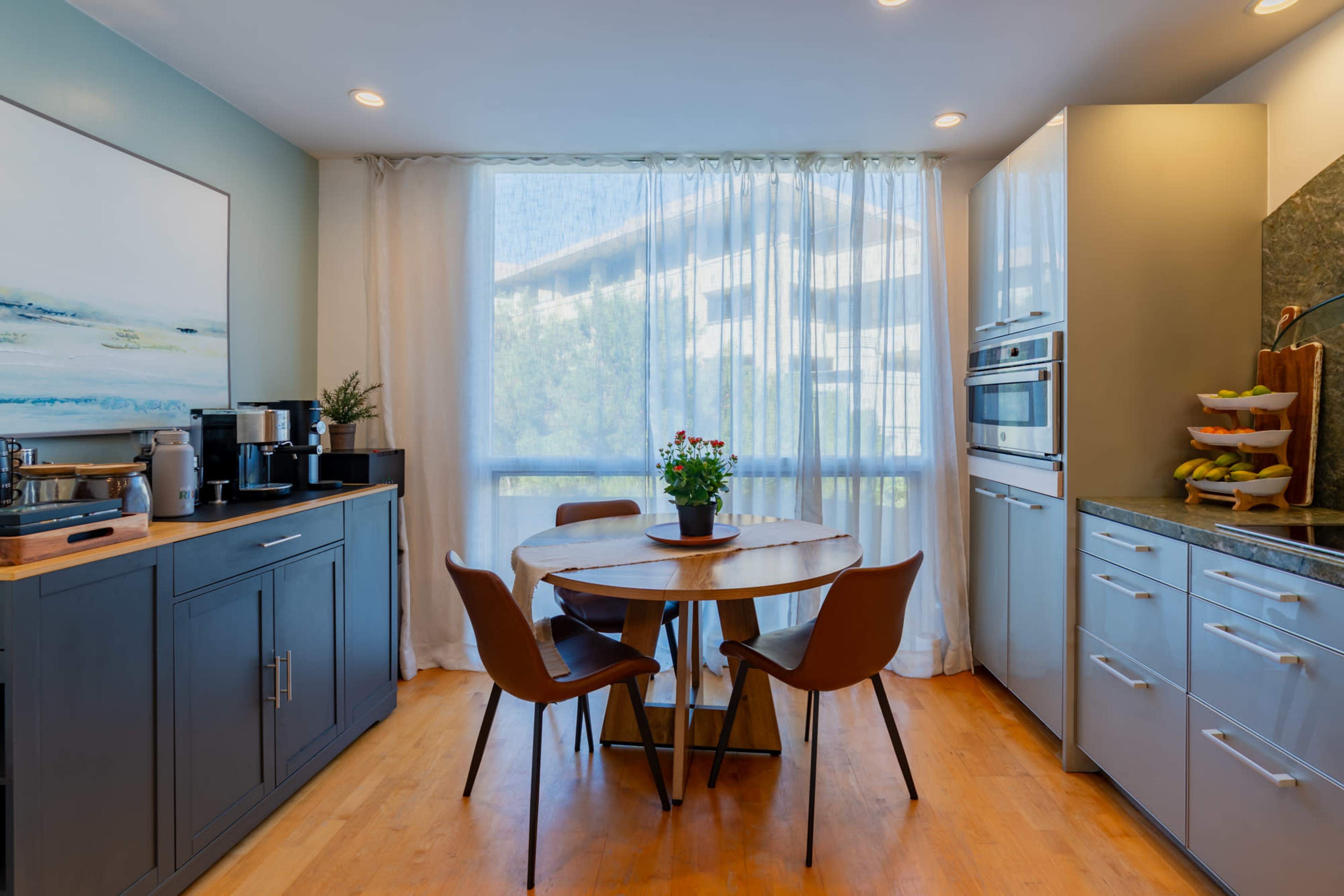 A bright kitchen features a round wooden table with four chairs, a coffee station, and modern appliances along the counter.