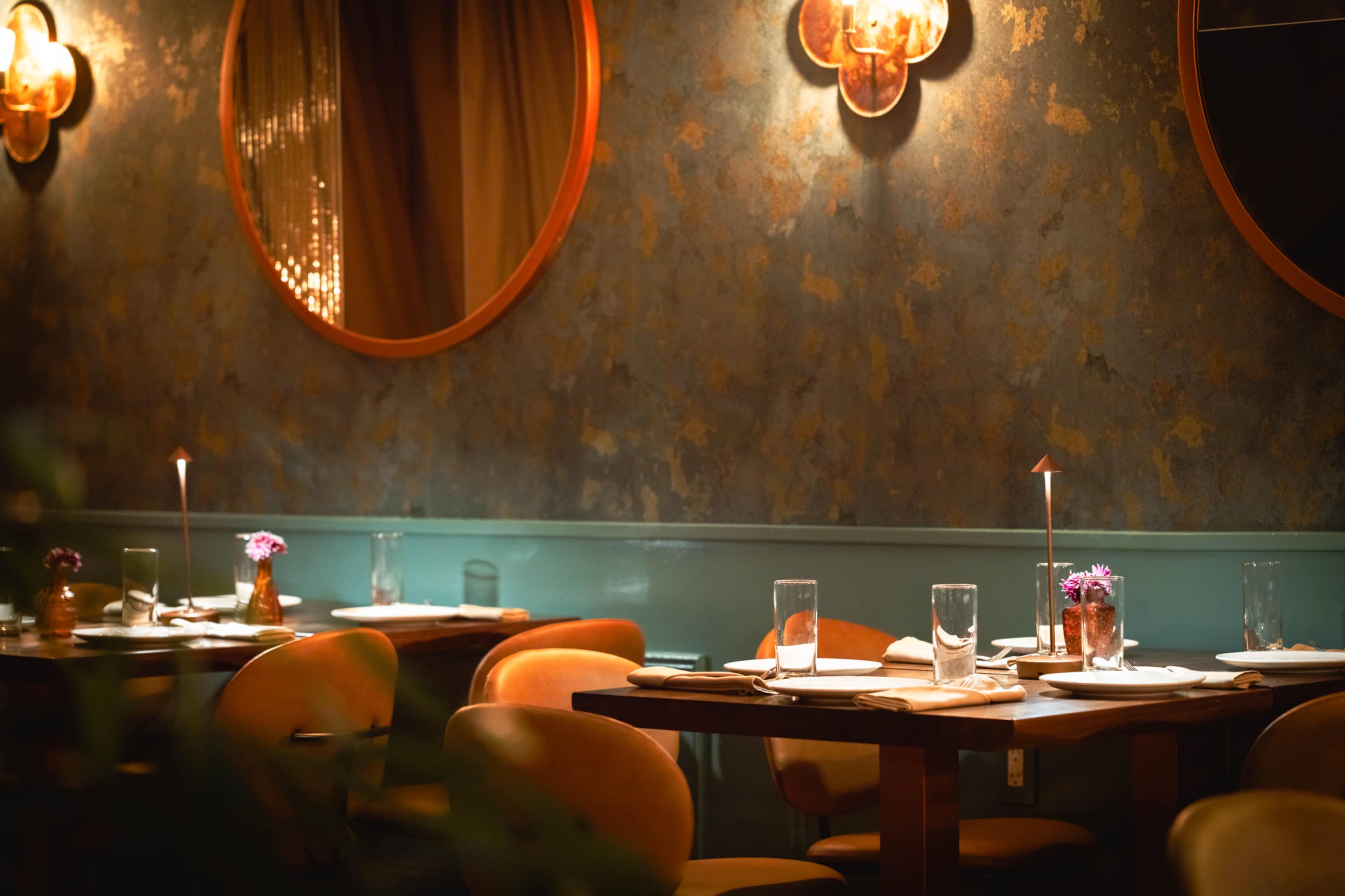 The image shows a dimly lit restaurant with circular mirrors on the walls, tables set with water glasses and small flower vases.