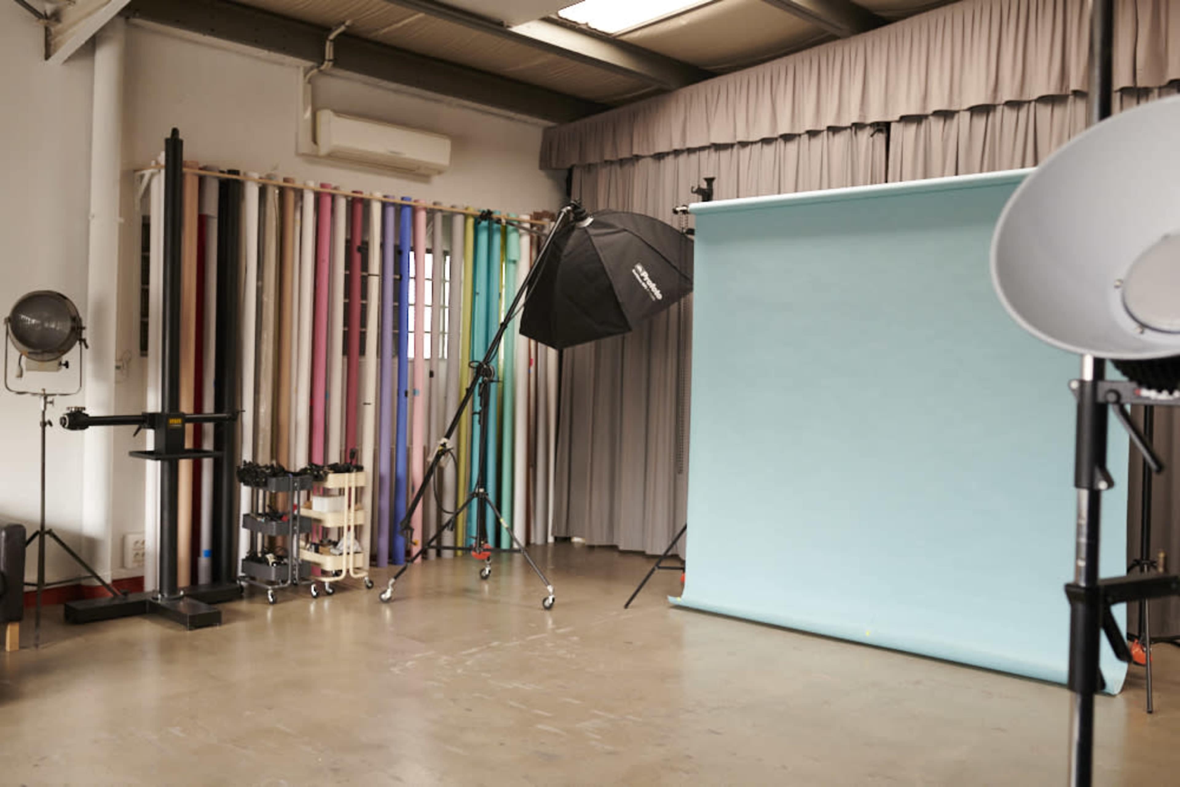 A photography studio features a light backdrop, lighting equipment, and colorful backdrop rolls arranged along a wall.