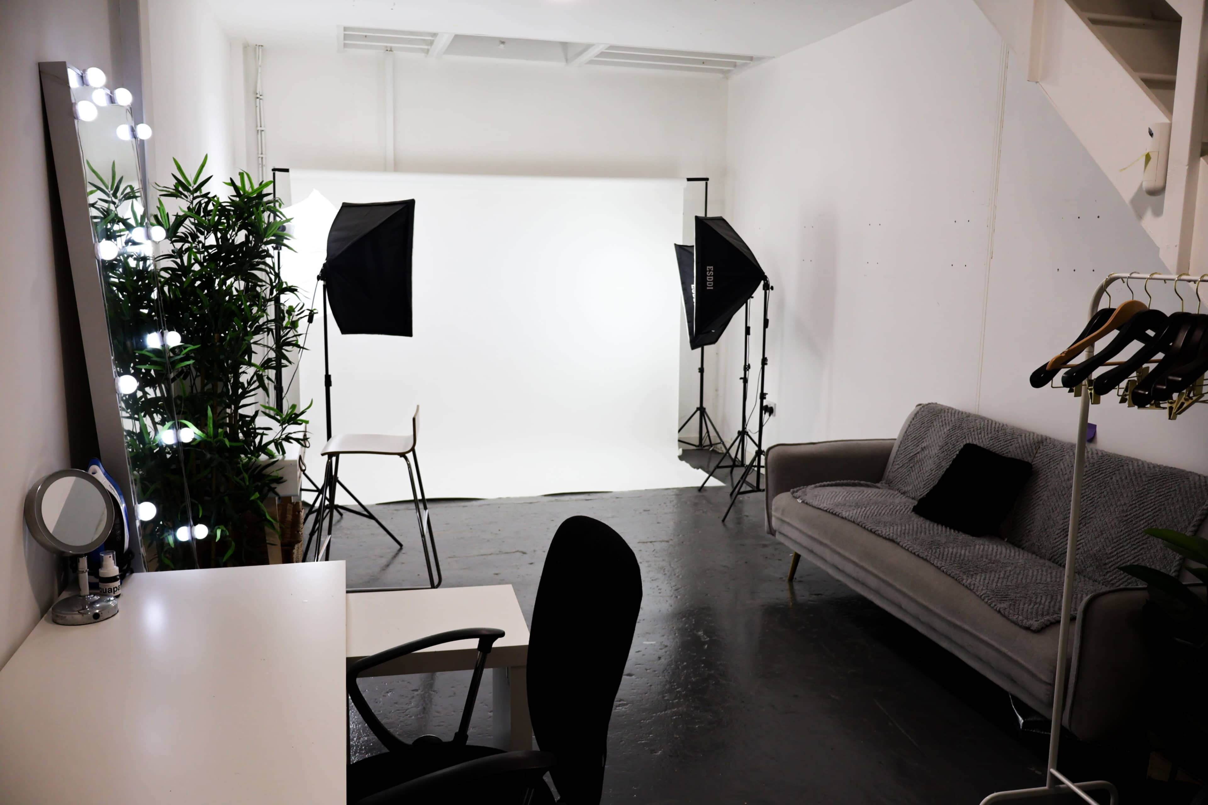 The image shows a minimalist photo studio setup with a white backdrop, two softboxes, a chair, a gray sofa, and a coat rack.