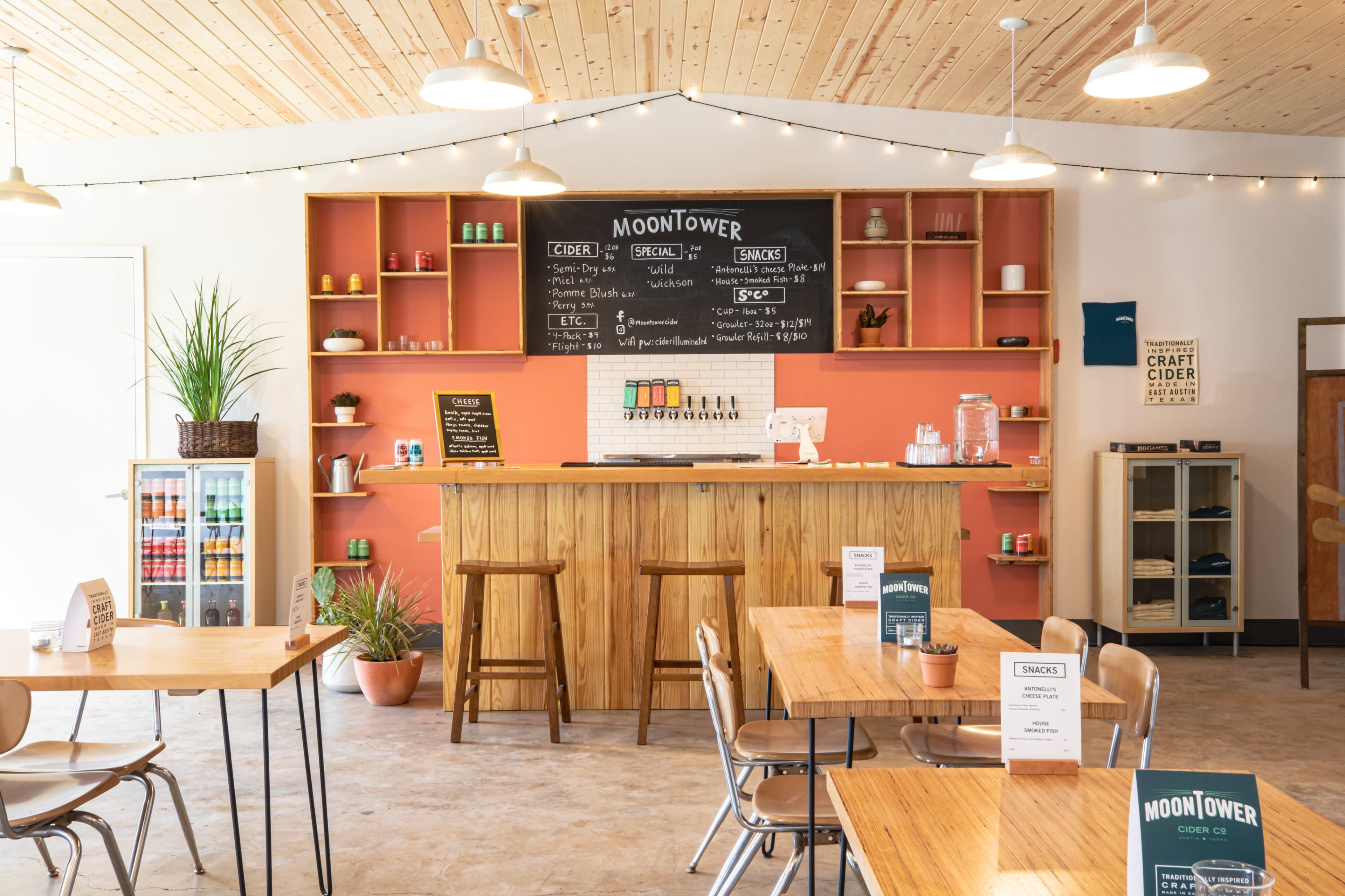 A cozy cider bar features a wooden counter, shelves with colorful bottles, and tables with menu signs under warm lighting.