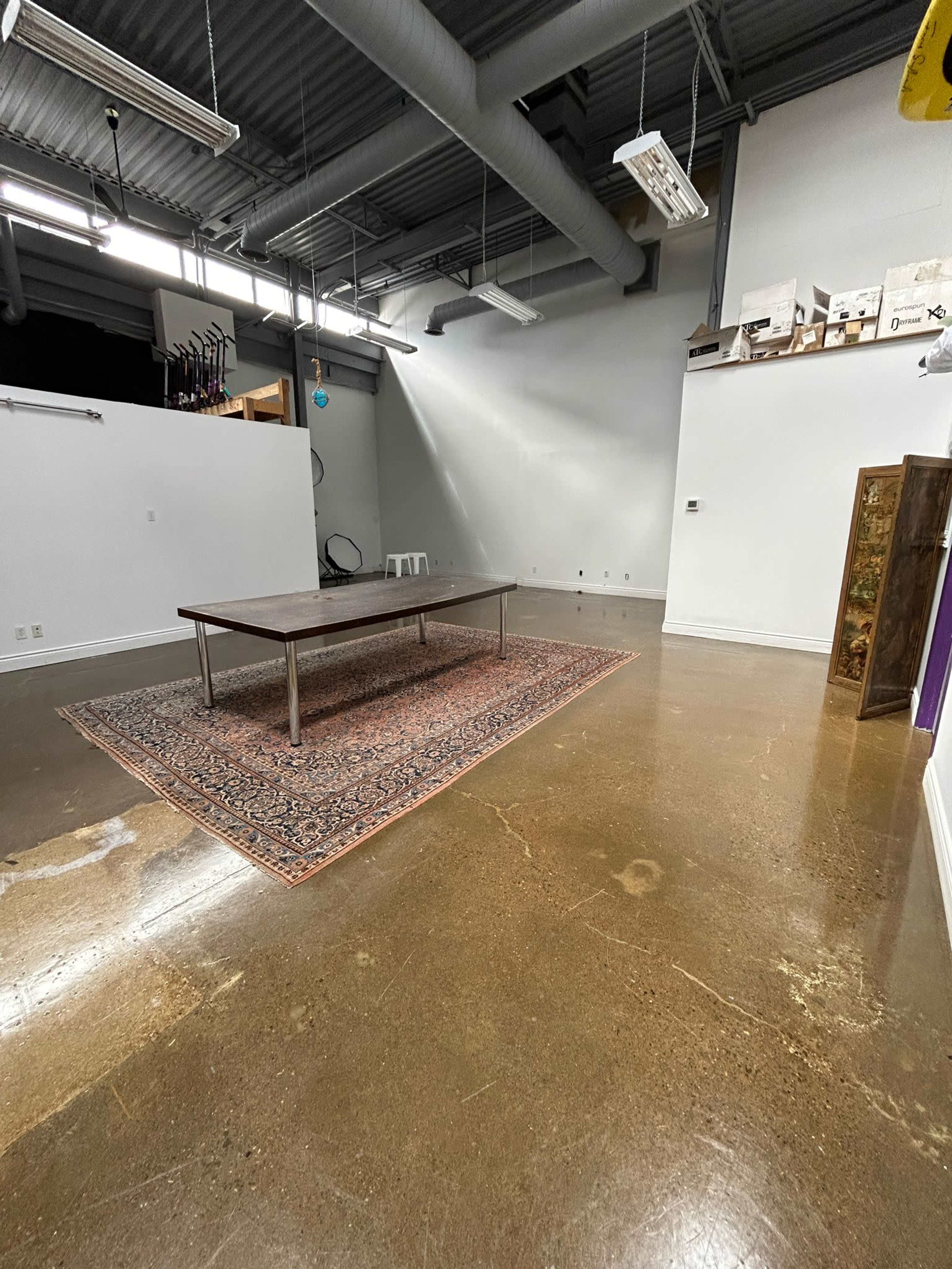 The image shows a spacious, empty room with a large wooden table placed on a patterned rug, and polished concrete floors.
