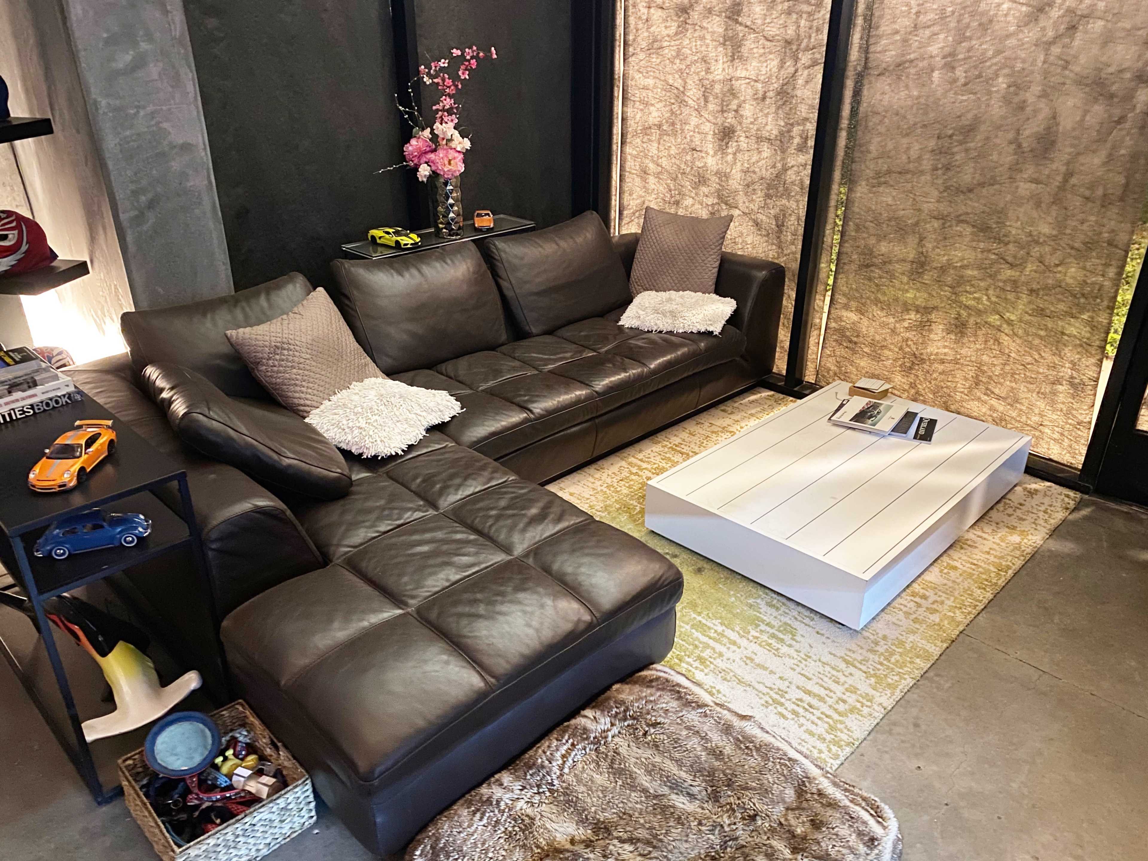 The image shows a modern living room featuring a dark leather sectional sofa, a white coffee table, and decorative accents on a gray wall with large windows covered by sheer curtains.