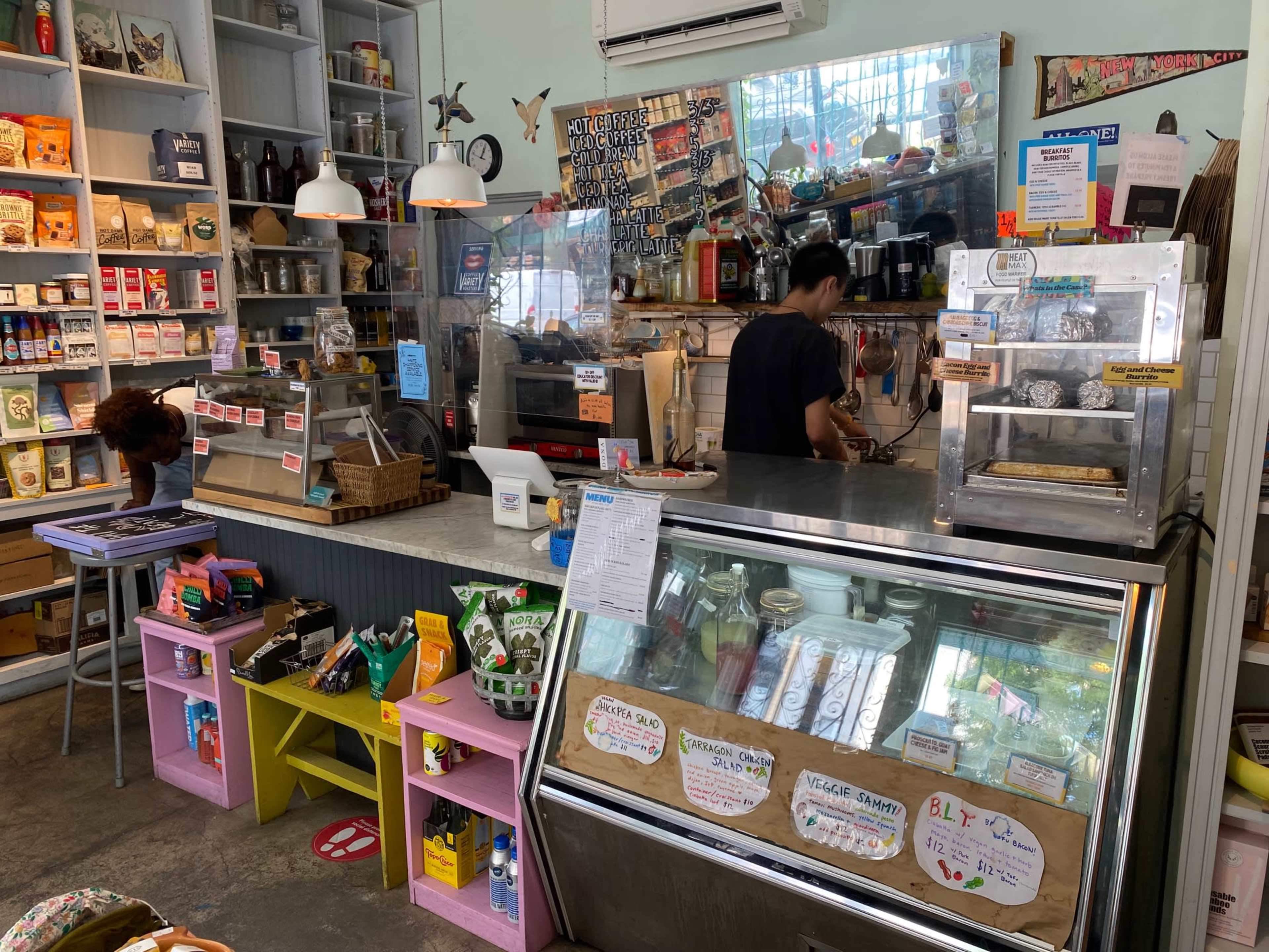 A café interior features a display counter with a glass case filled with pastries and snacks, while staff members prepare food and drinks behind the counter.