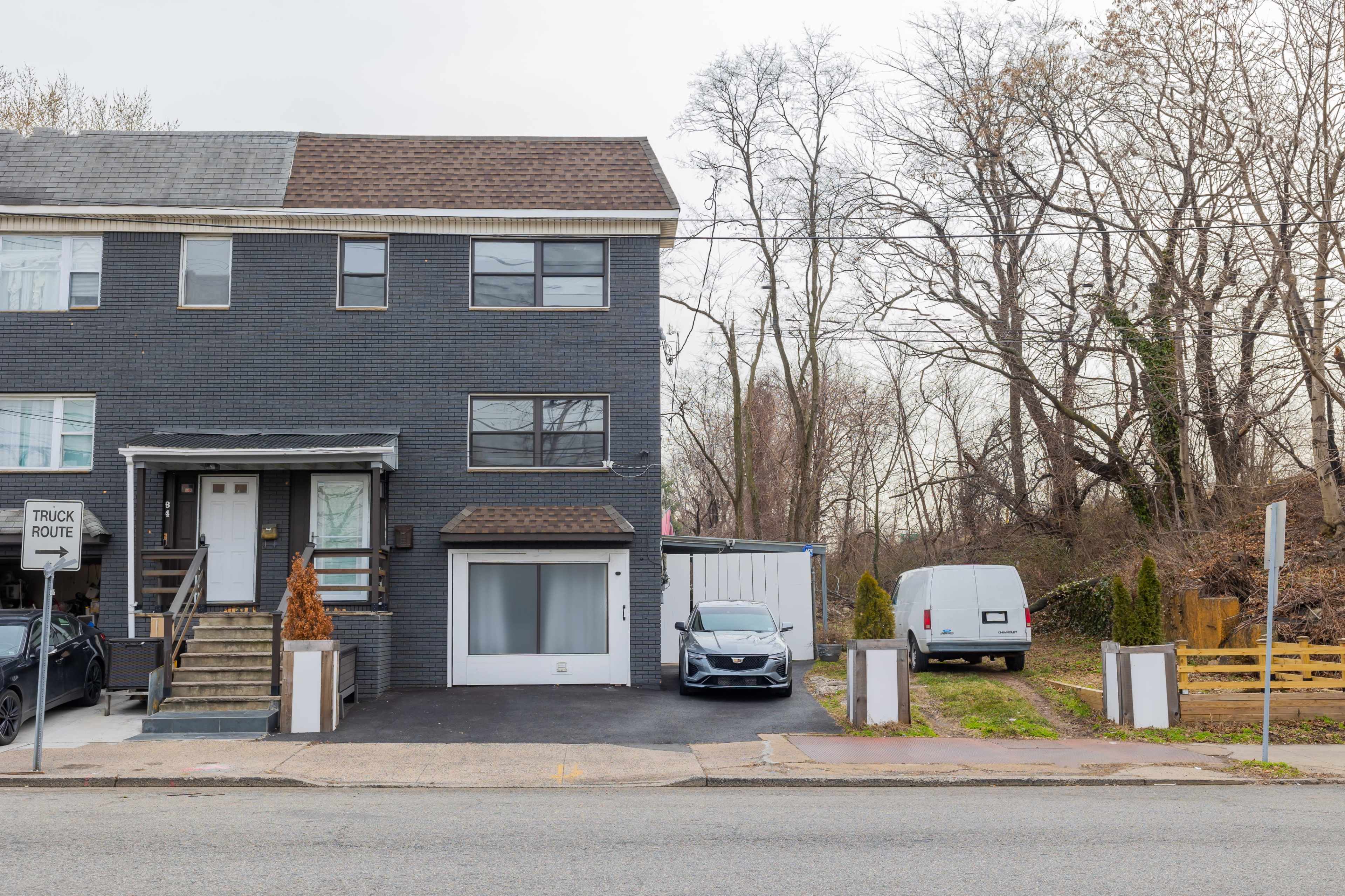 A two-story brick house with a gray facade and a garage is situated next to a white van and a vacant lot lined with trees.