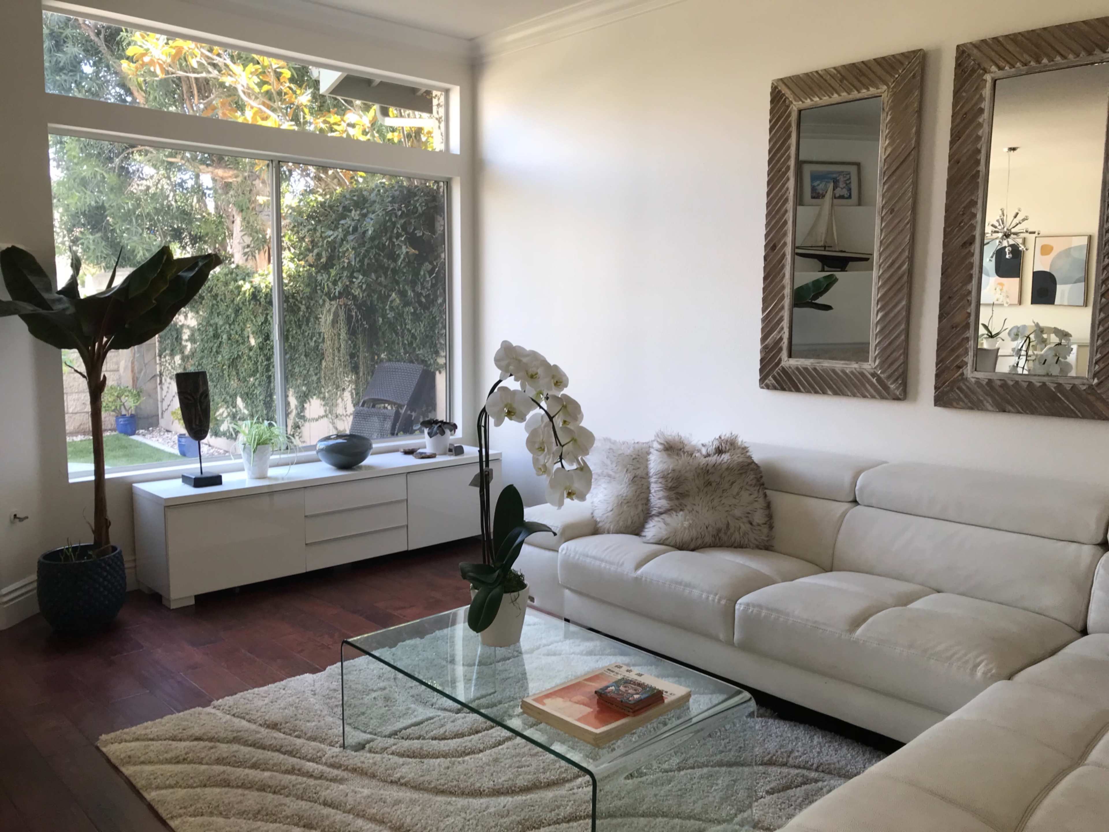 A bright living room features a white sectional sofa, a glass coffee table, and large windows that provide a view of an outdoor garden.