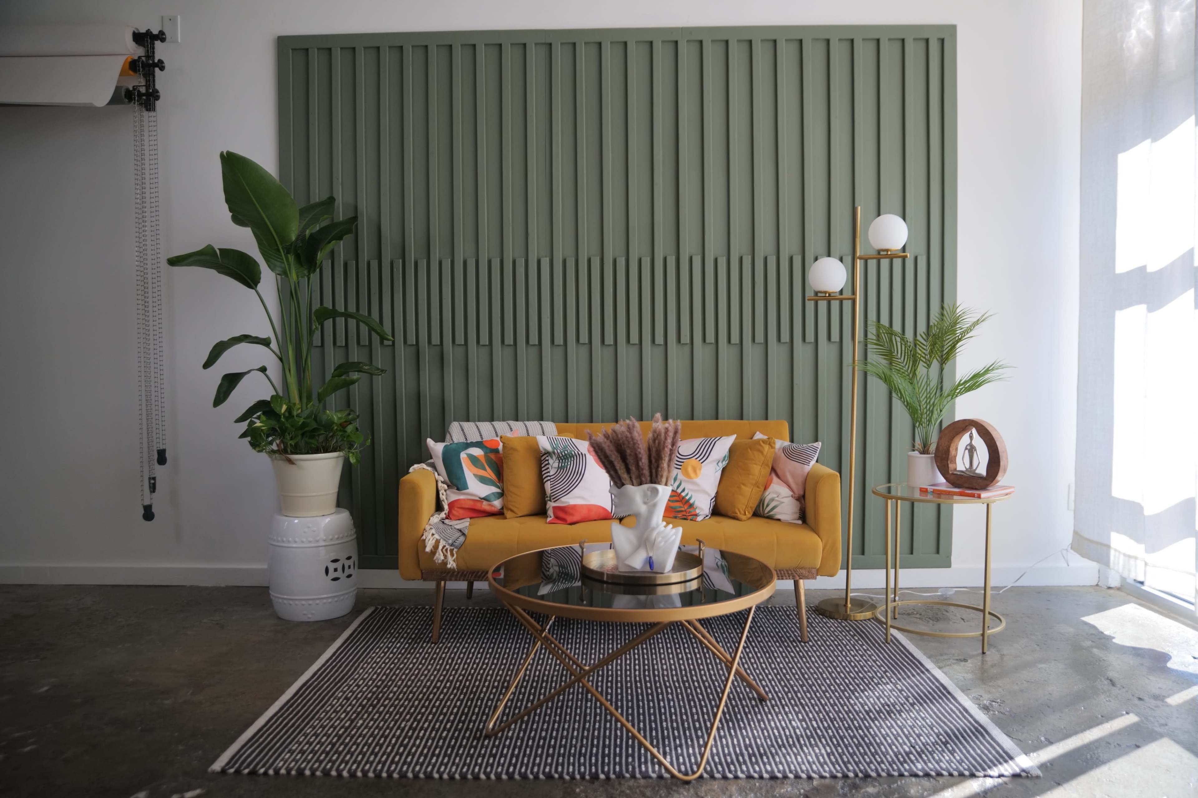 A yellow sofa with decorative pillows is positioned in front of a green vertical panel wall, accompanied by a coffee table, indoor plants, and a patterned rug.